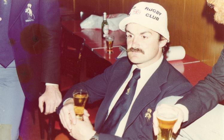 A man wearing a rugby club hat and blazer holding a glass of beer, sitting in a casual setting.