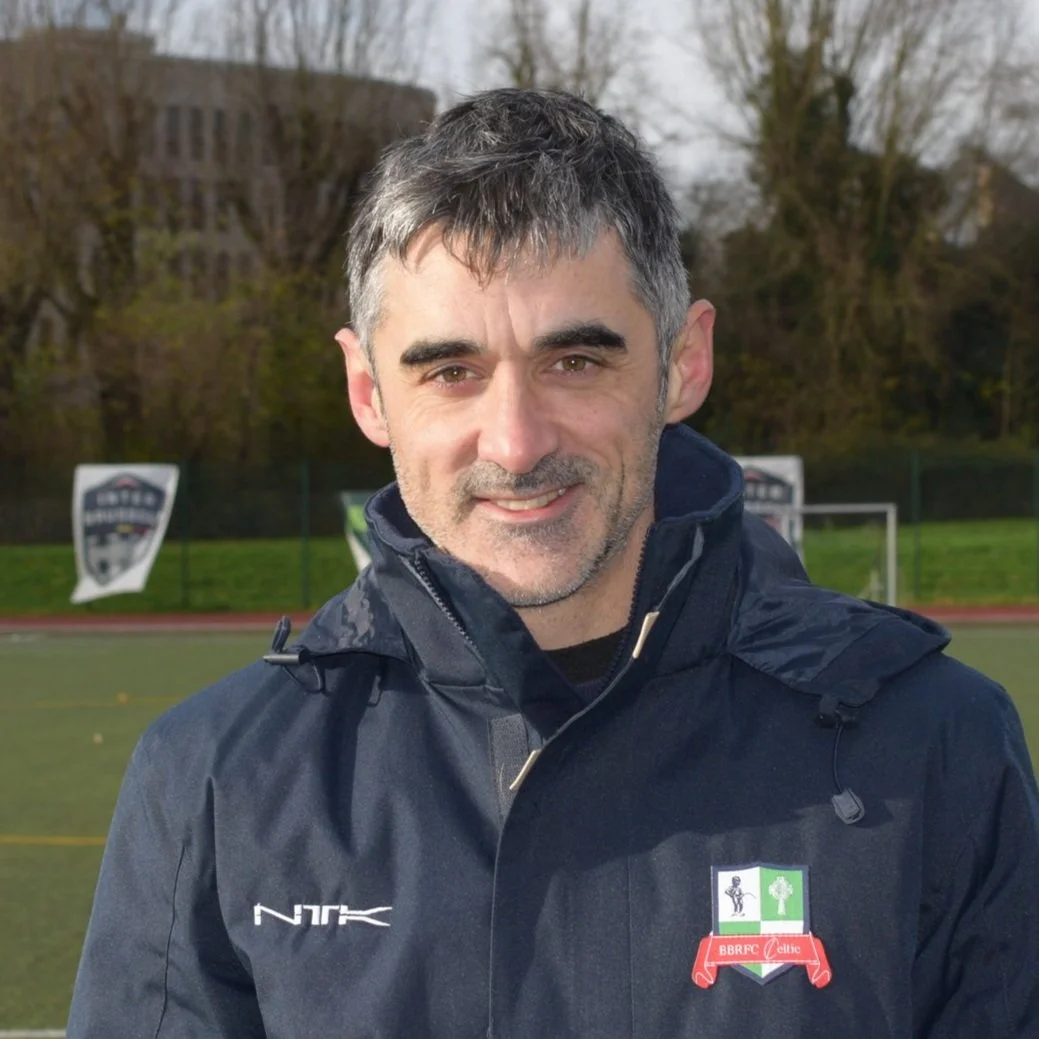 A man wearing a dark jacket on a sports field with banners in the background.