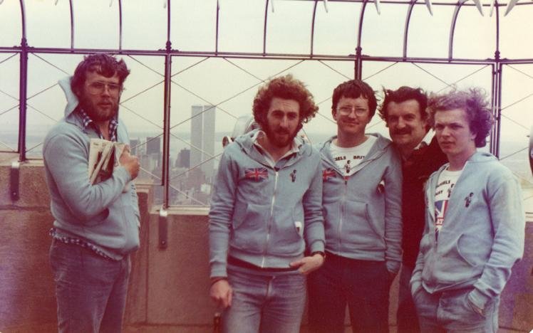 Group of five men posing at an elevated observation deck with cityscape background, wearing casual clothing, including matching light blue hoodies.
