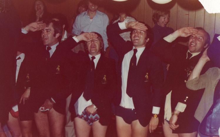 Four men in jackets and ties without pants saluting indoors with onlookers in the background.