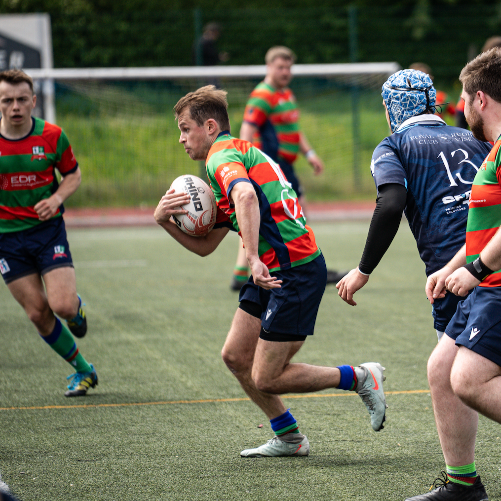 Rugby players in action during a match, wearing blue and red uniforms, competing for the ball on a grassy field.