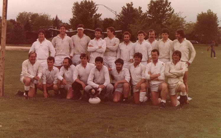 A rugby team posing for a group photo on a grass field, wearing white uniforms, surrounded by trees.