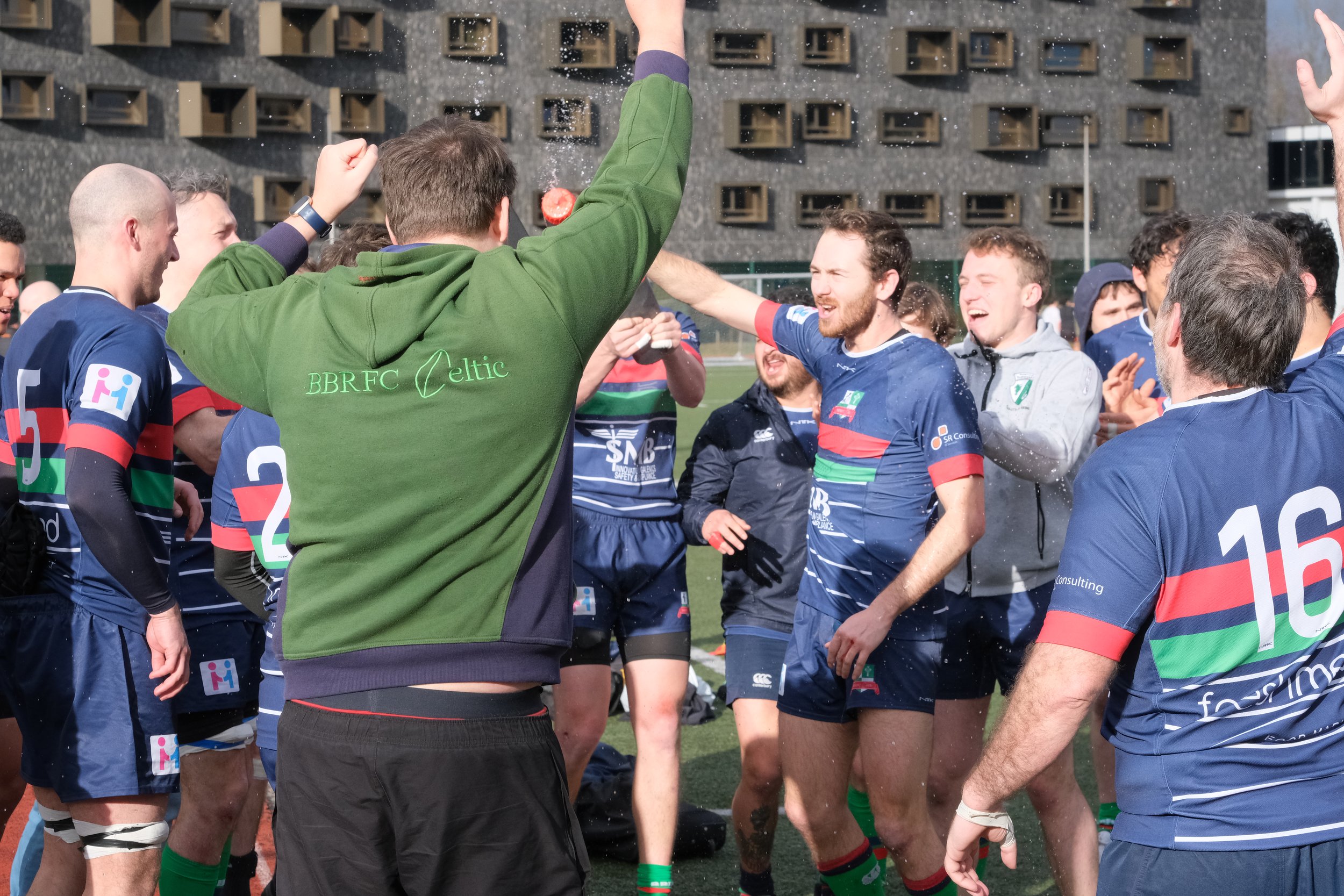 Group of rugby players celebrating, wearing blue jerseys with colorful stripes, outdoors on a field.