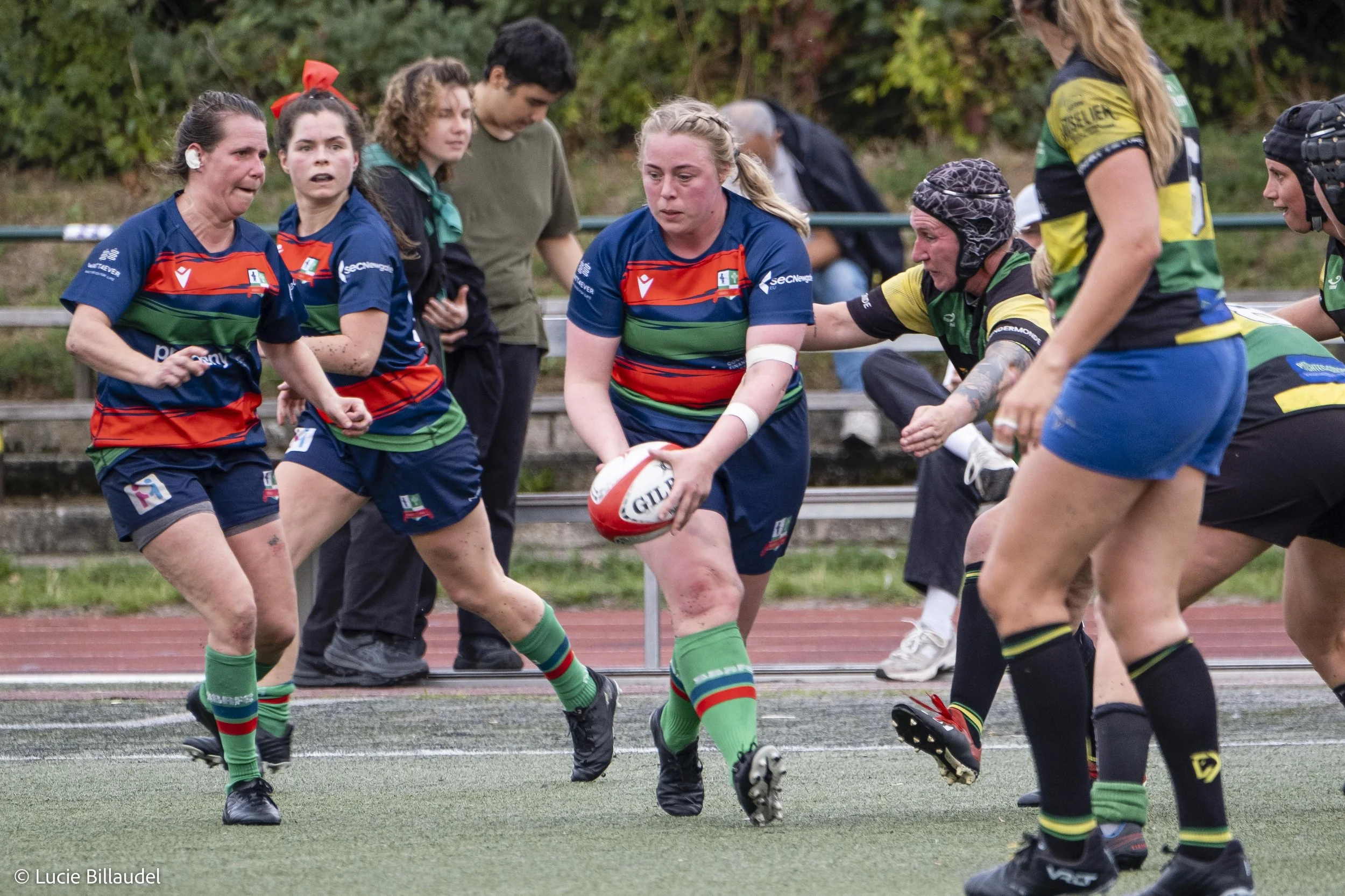 Women playing rugby on a field, one running with the ball in a green and blue jersey, others in pursuit.