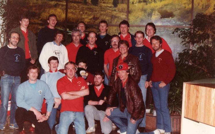 Group of men posed indoors, wearing casual sweaters and jackets, in front of a nature mural.