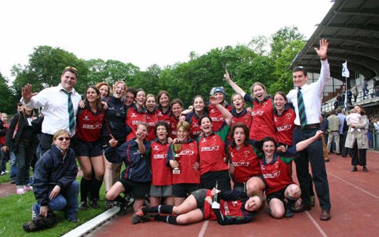 Women's rugby team celebrating victory with a trophy on a sports field.