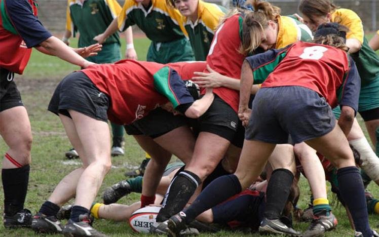 Women playing rugby in a scrum, wearing red and green uniforms, on a grassy field.