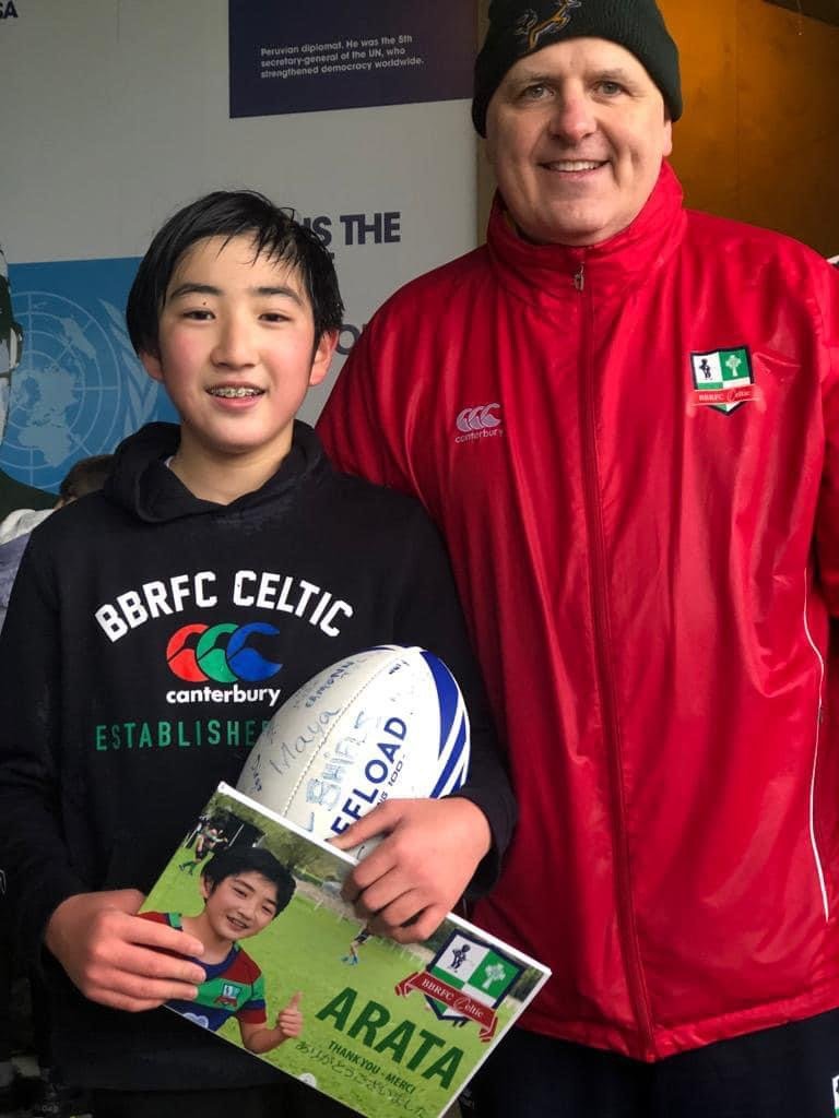 Two people posing, one holding a rugby ball and a booklet. The younger one is wearing a black hoodie with "BBRFC Celtic" and "Canterbury" logos, the older one in a red jacket with a rugby logo.