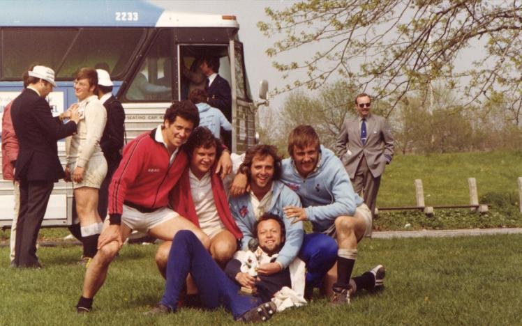 Group of men posing near a bus with a dog, wearing sports attire in a grassy area with trees.