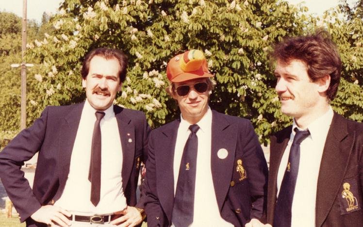 Three men wearing suits and ties, one with a novelty hat, outdoors with trees in the background.