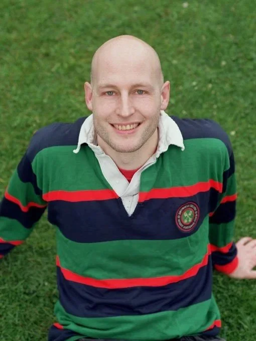 Person standing on sports field at dusk, wearing a jacket, smiling with arms crossed.