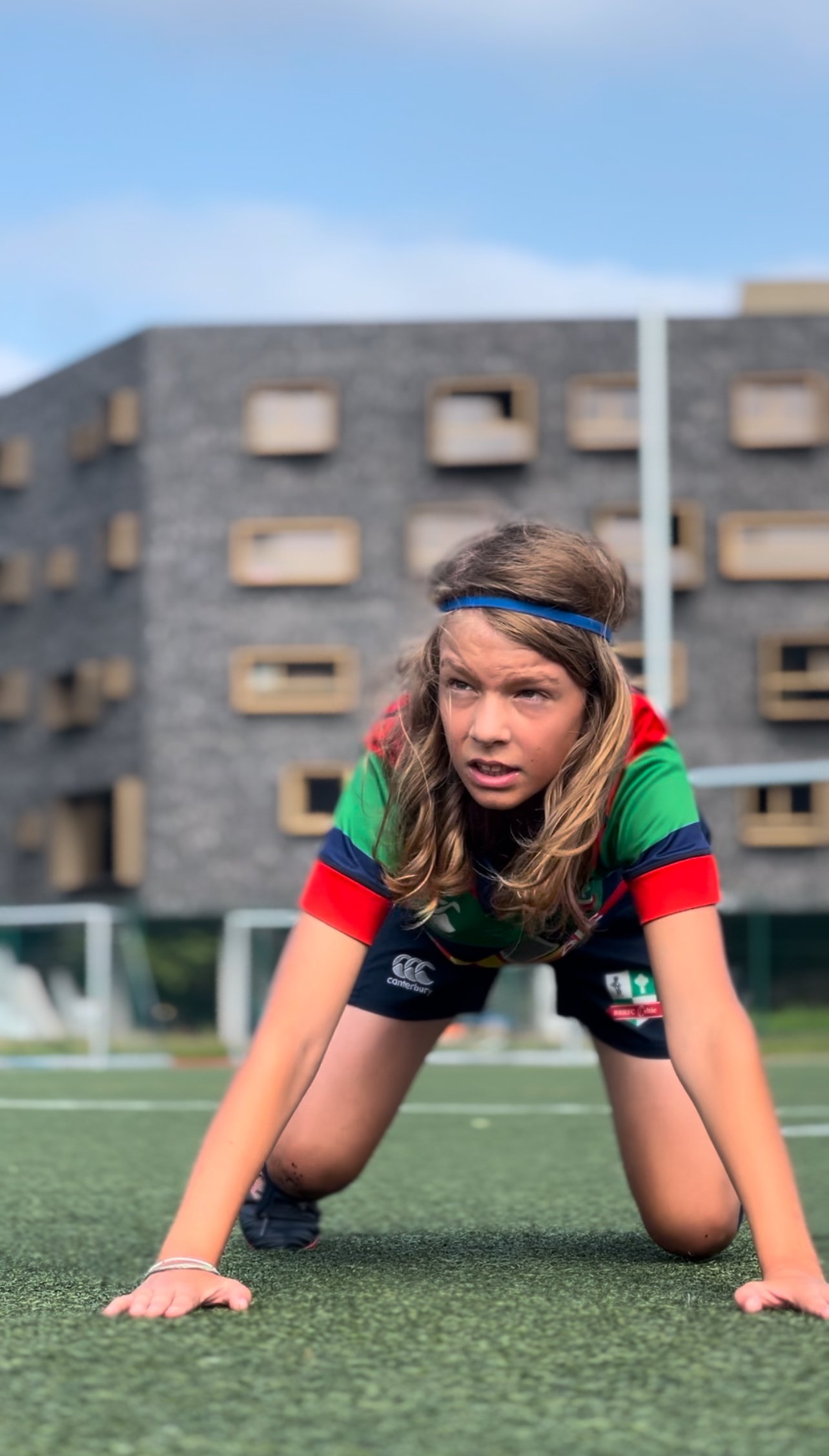 Young athlete in a starting position on a grass field, wearing a colorful sports jersey and headband, with a modern building in the background.