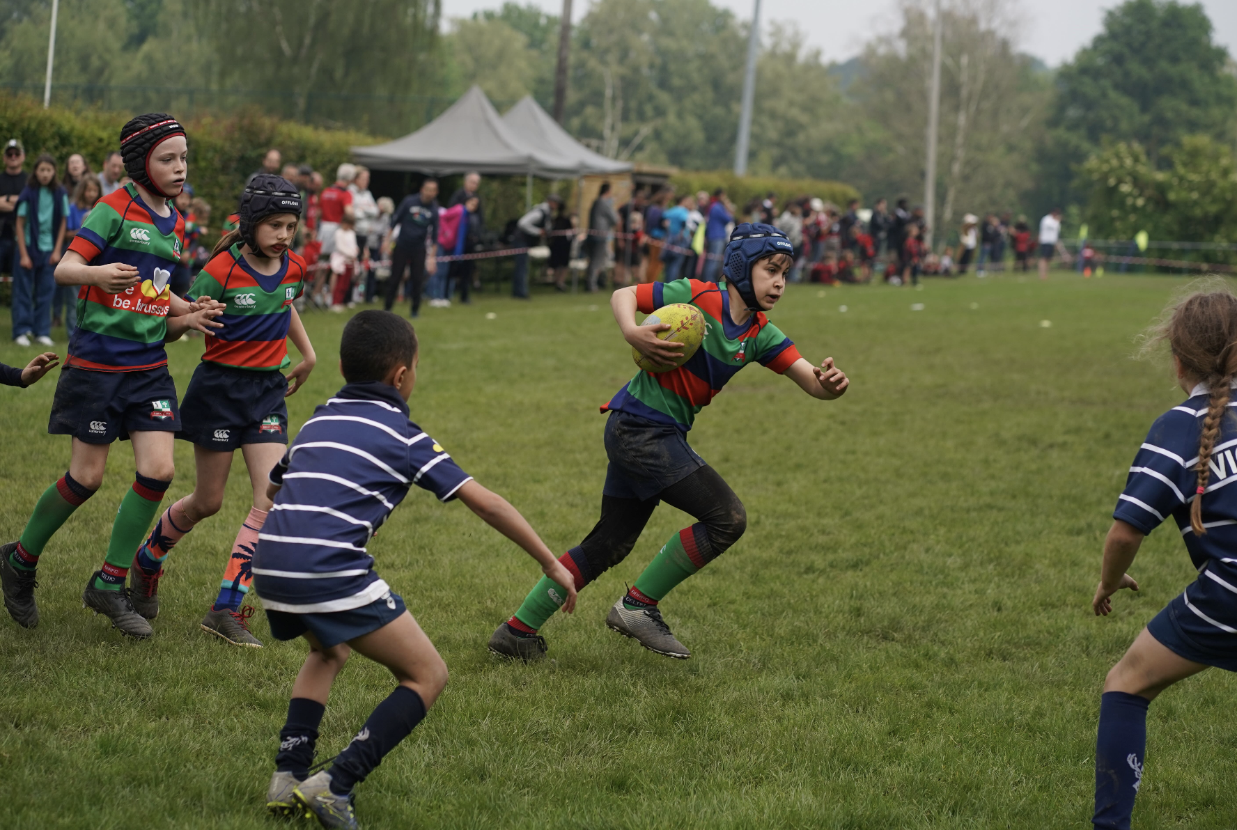 Children playing rugby, outdoor field, teams in colorful jerseys, spectators in background.