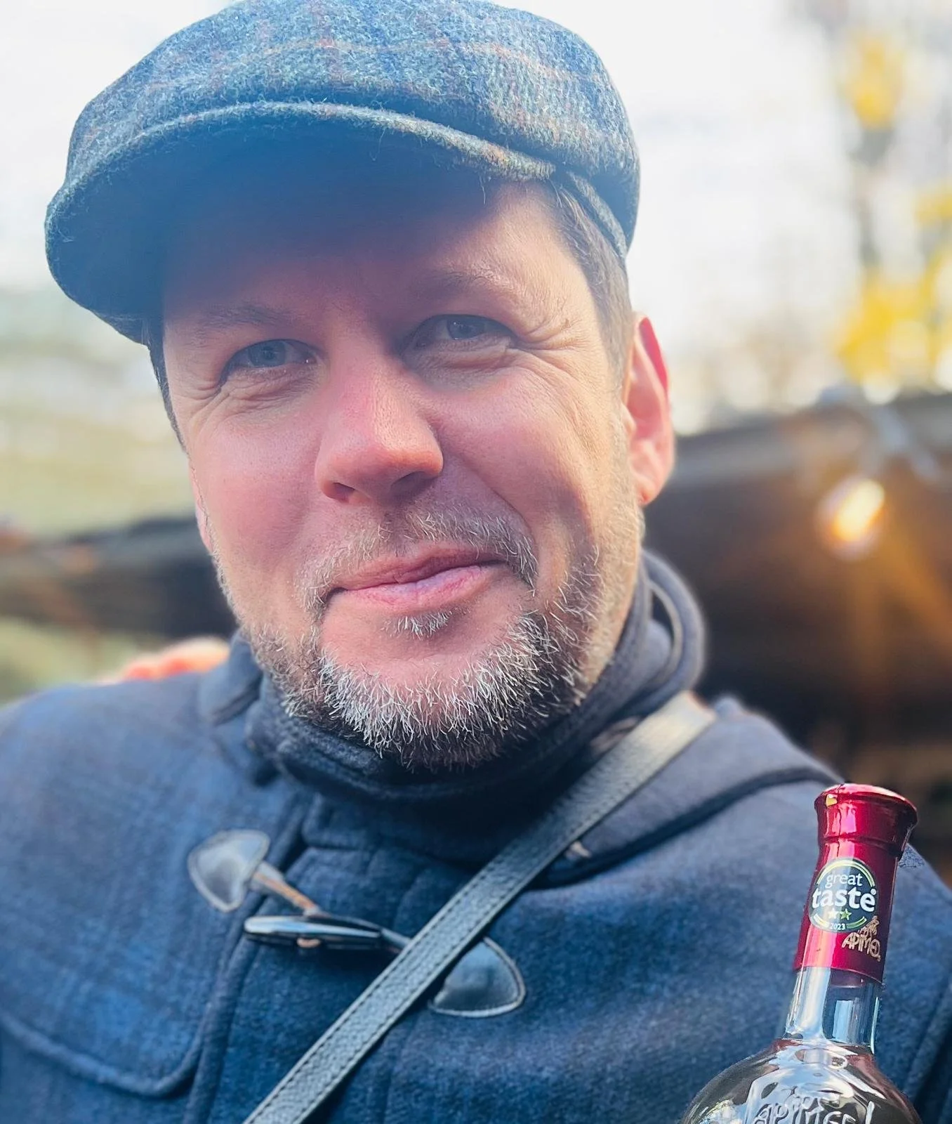 Man in blue jacket and flat cap holding a bottle outdoors.