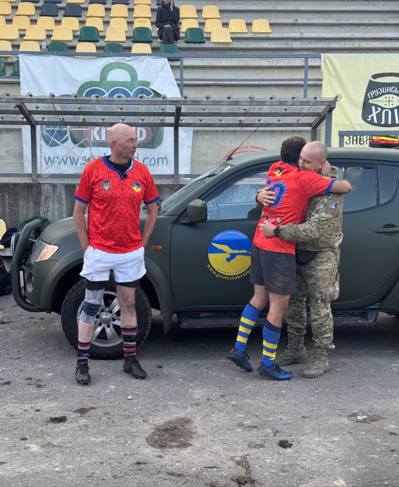 Trois hommes en tenue de sport et en uniforme militaire près d'un véhicule avec un logo, debout devant des sièges de stade.