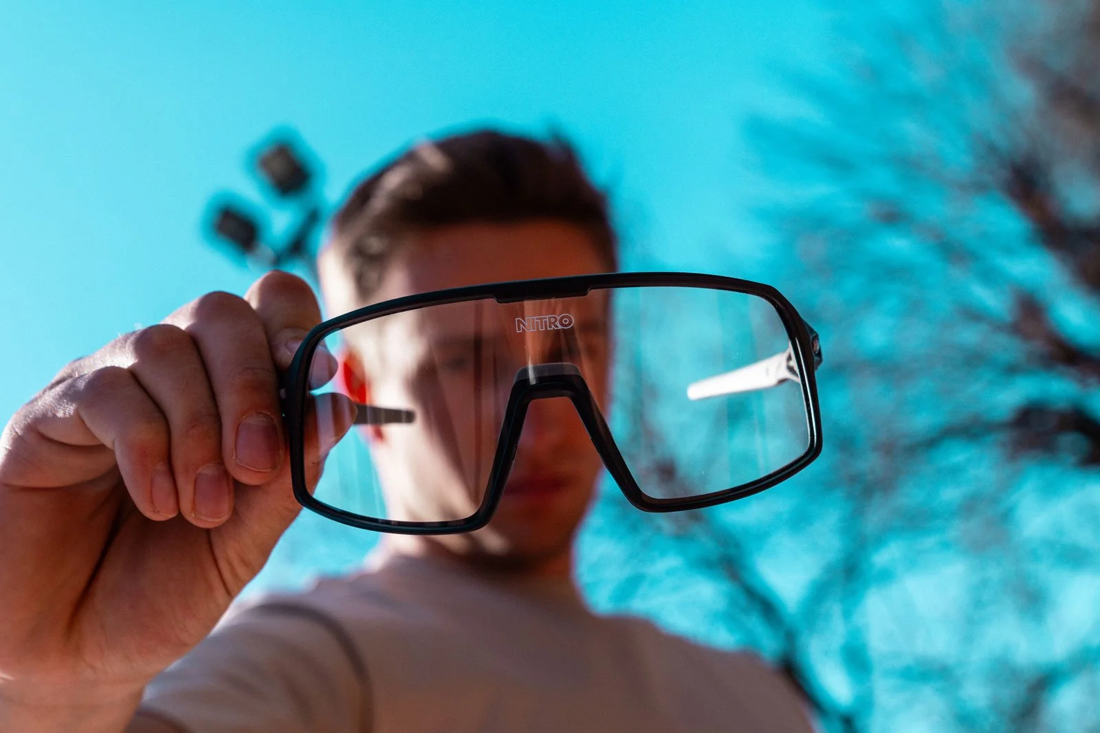 Un ragazzo tiene una paio di occhiali da sole davanti alla fotocamera, con sfondo blu e alberi.