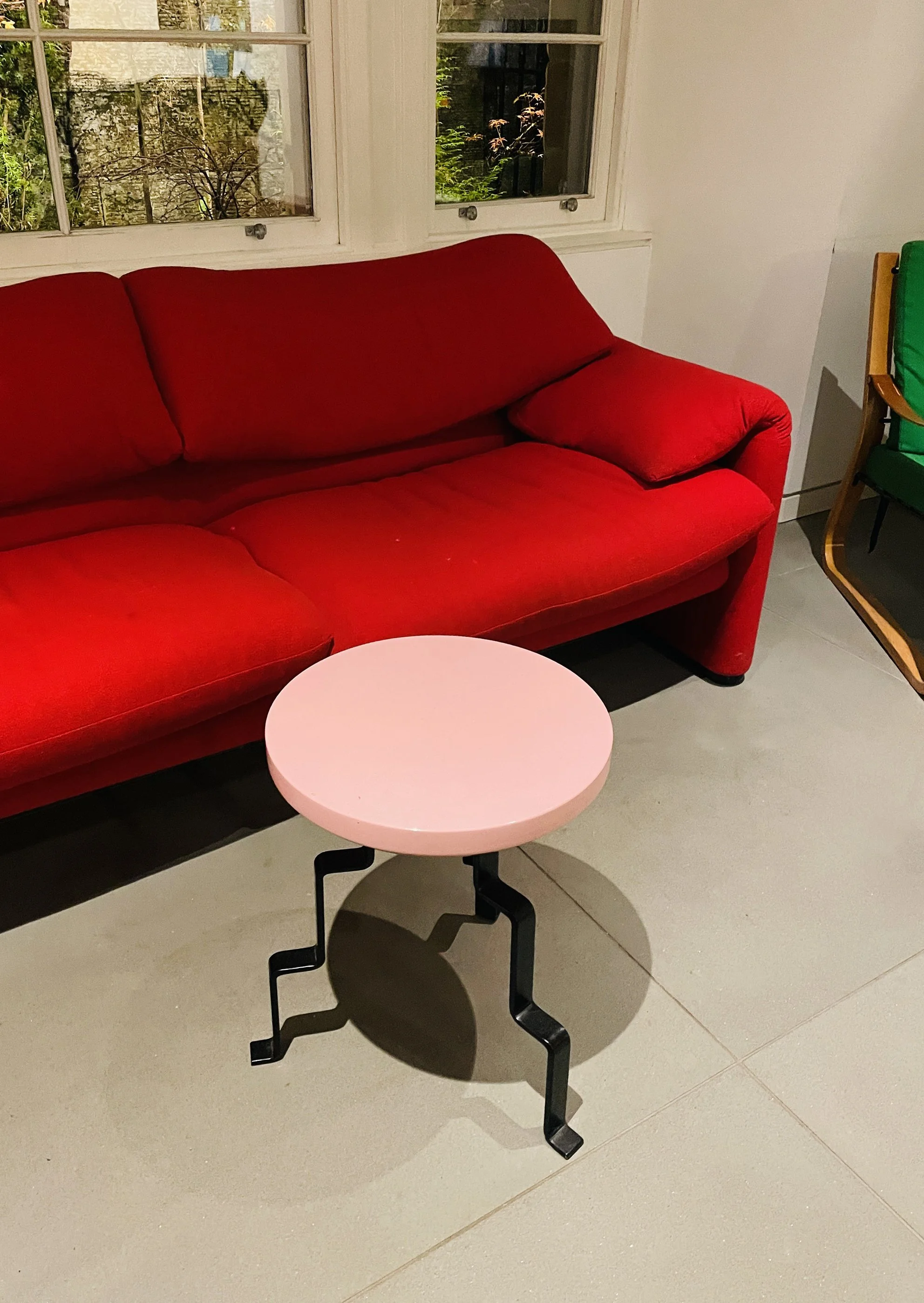 A red sofa with a matching red cushion, a pink round side table with black zigzag legs, white walls, and windows showing trees outside.