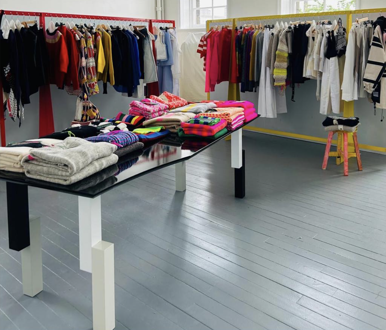 Clothing store with colorful sweaters, shirts, and scarves displayed on racks and a black table with folded items, set against a white wall and gray wooden floor.