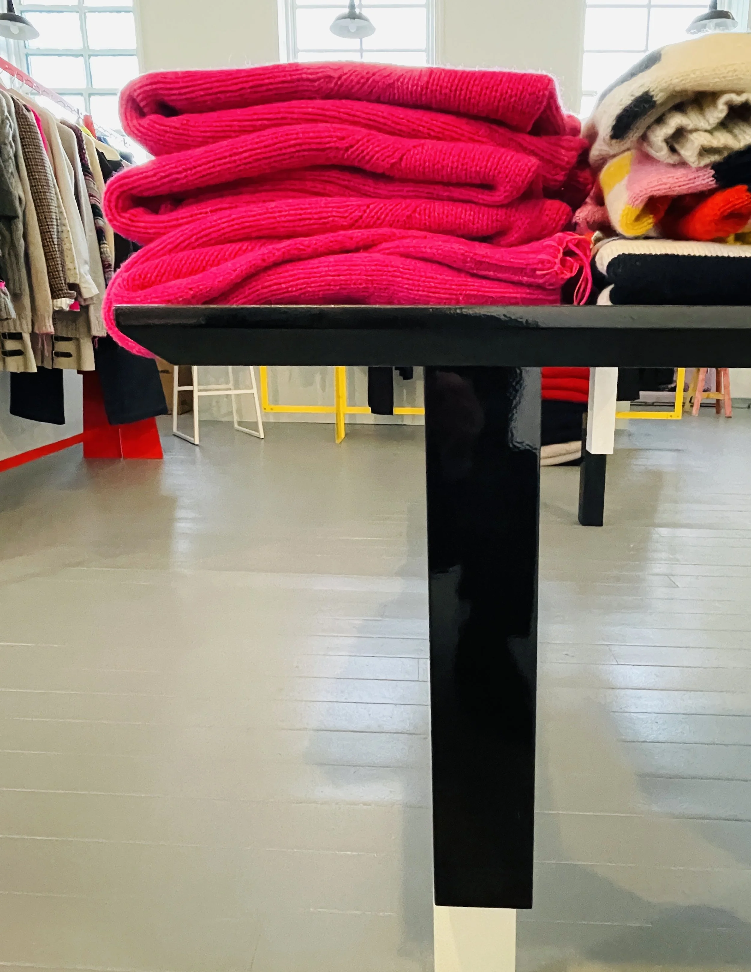 Stack of bright pink folded sweaters on a black table in a clothing store, with other clothing items and racks visible in the background.
