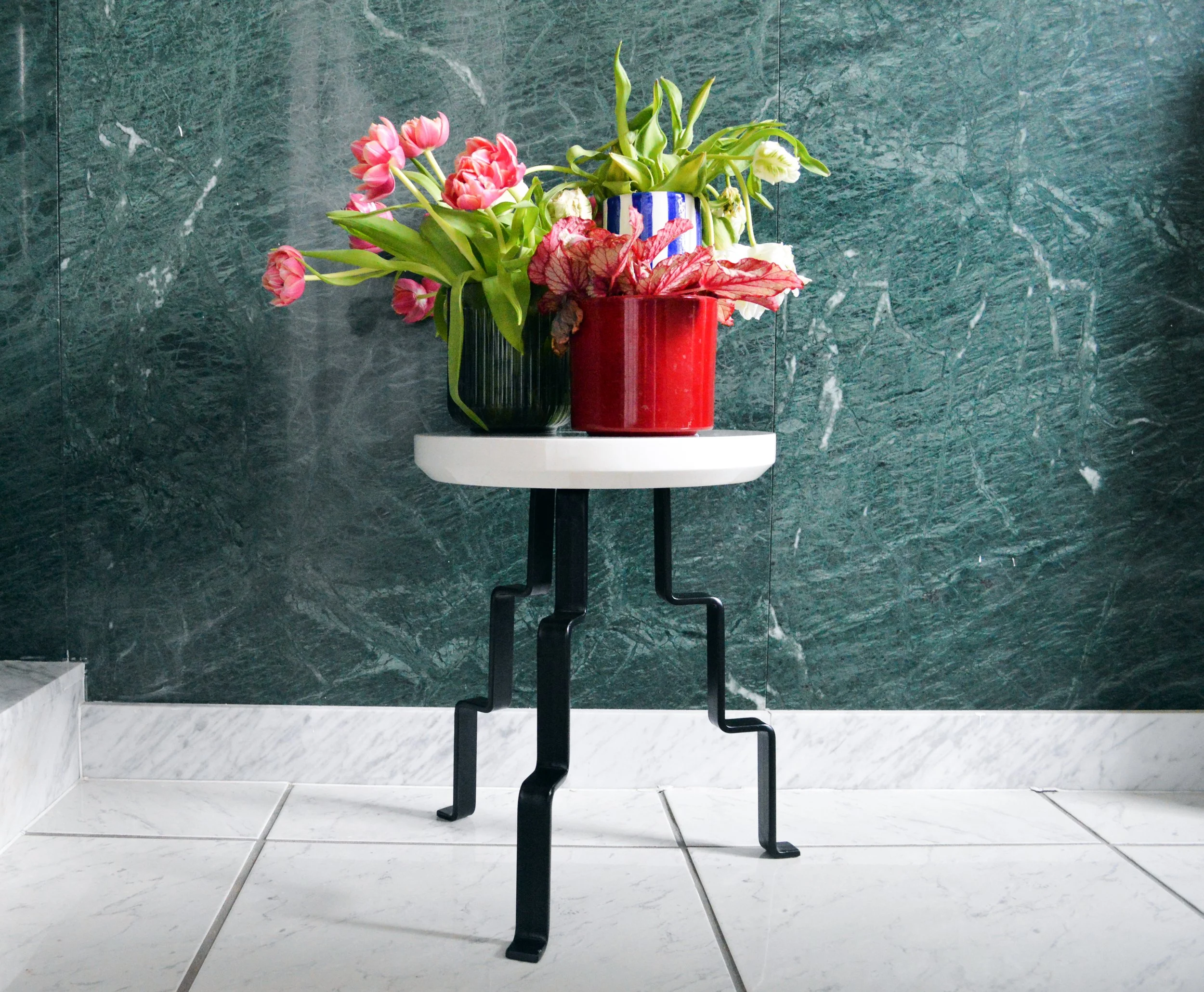 A small round table with black metal legs and a white top, holding three flower vases with tulips, placed against a green marble wall and white tiled floor.