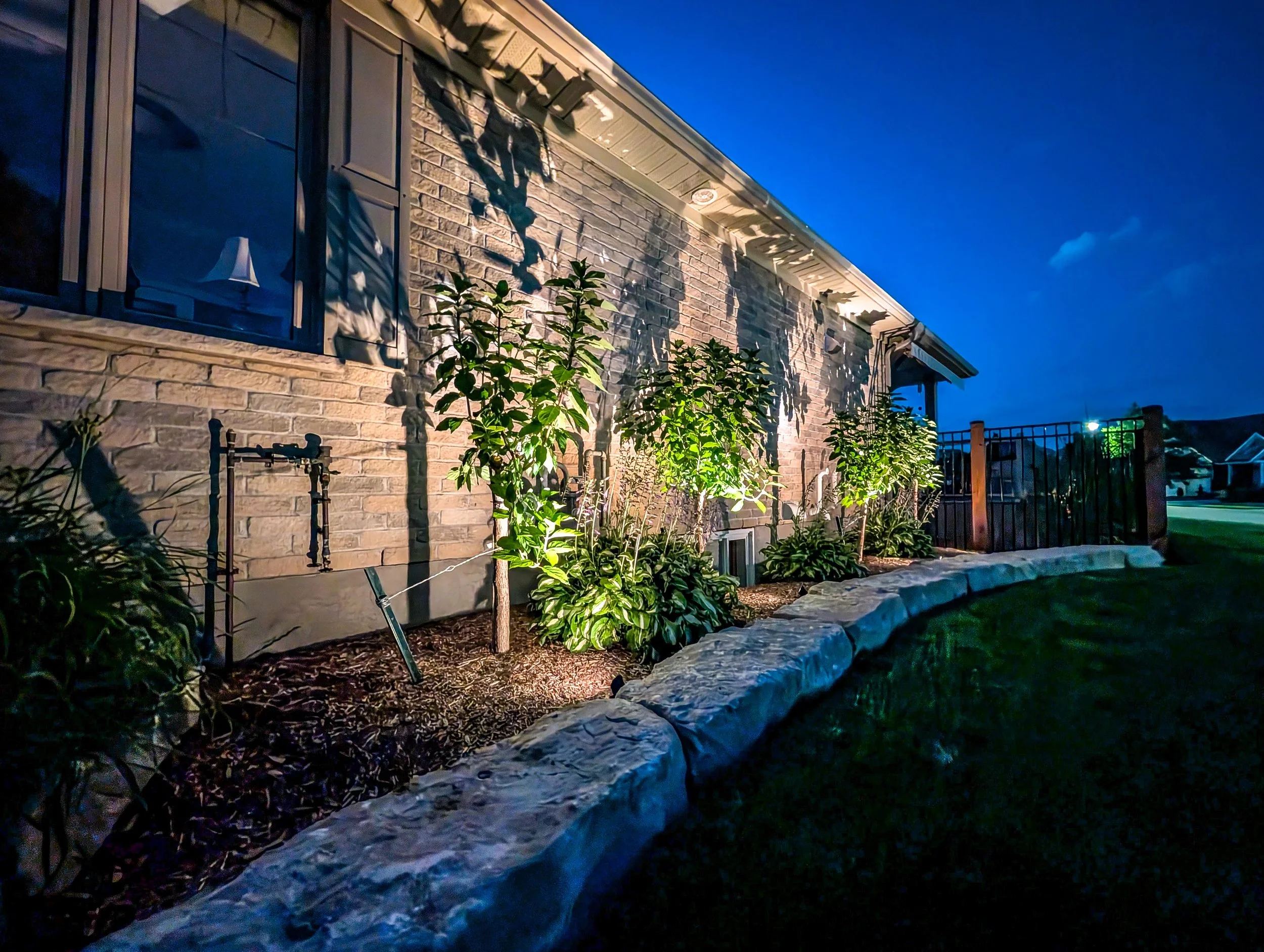 A view of the side of a brick house at dusk with a blue sky. Shadows of a tree are cast on the house wall, and there are green plants in the garden illuminated by outdoor lighting.