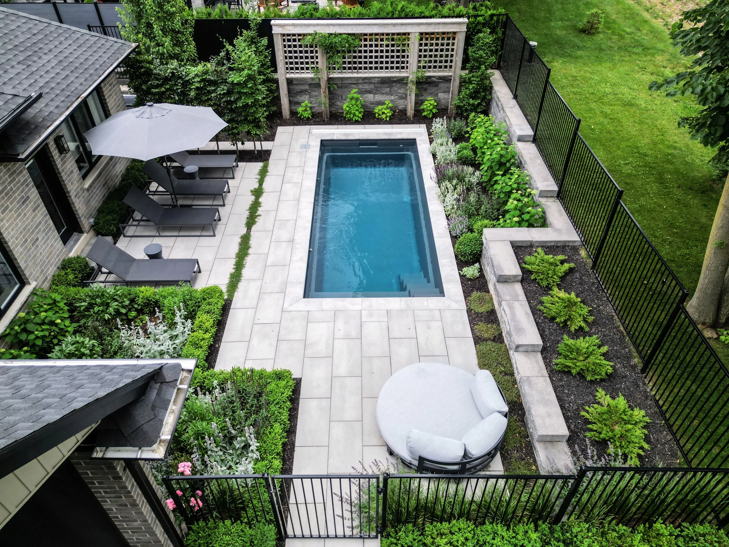 A backyard patio featuring a rectangular swimming pool, surrounded by light-colored tiles, with lounge chairs and umbrellas on one side, a small round seating area with cushions, lush greenery, flower beds, and a black metal fence enclosing the area.