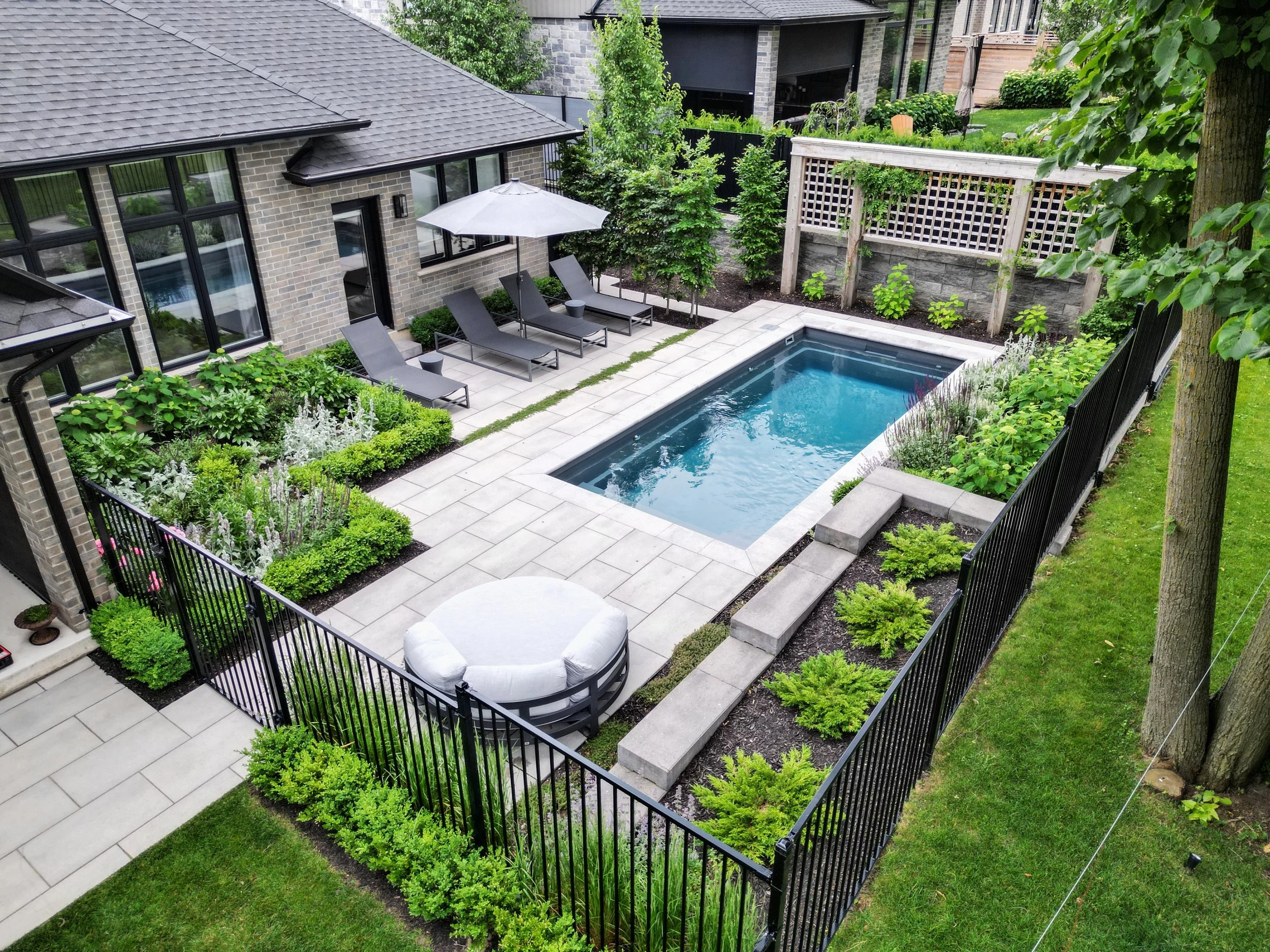 A backyard with a swimming pool, lounge chairs, umbrella, and landscaped garden surrounded by a black metal fence.
