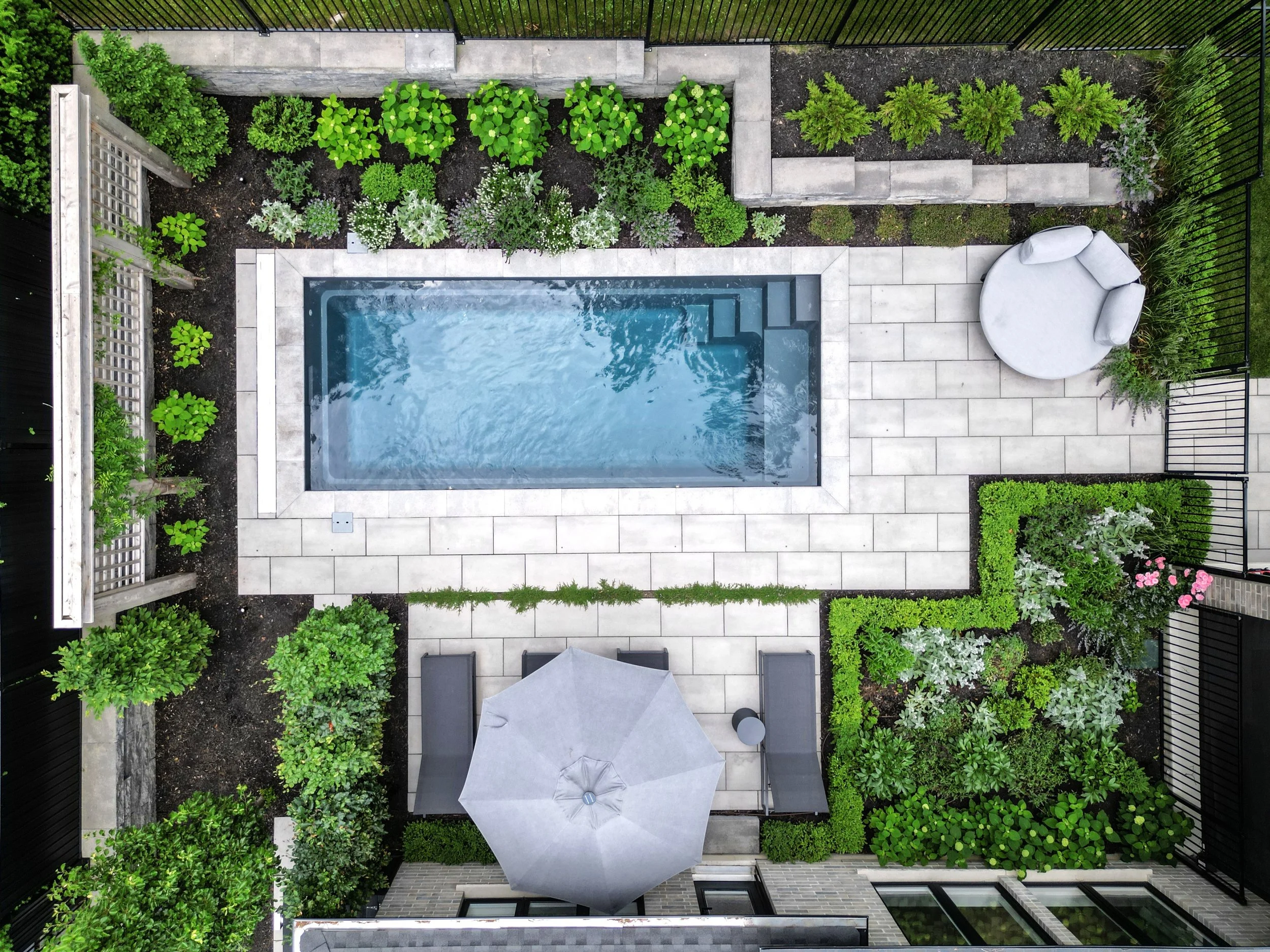 Aerial view of a backyard with a small rectangular swimming pool, surrounded by tiled patio, garden beds with various green plants, a sitting area with an umbrella, and a lounge chair.