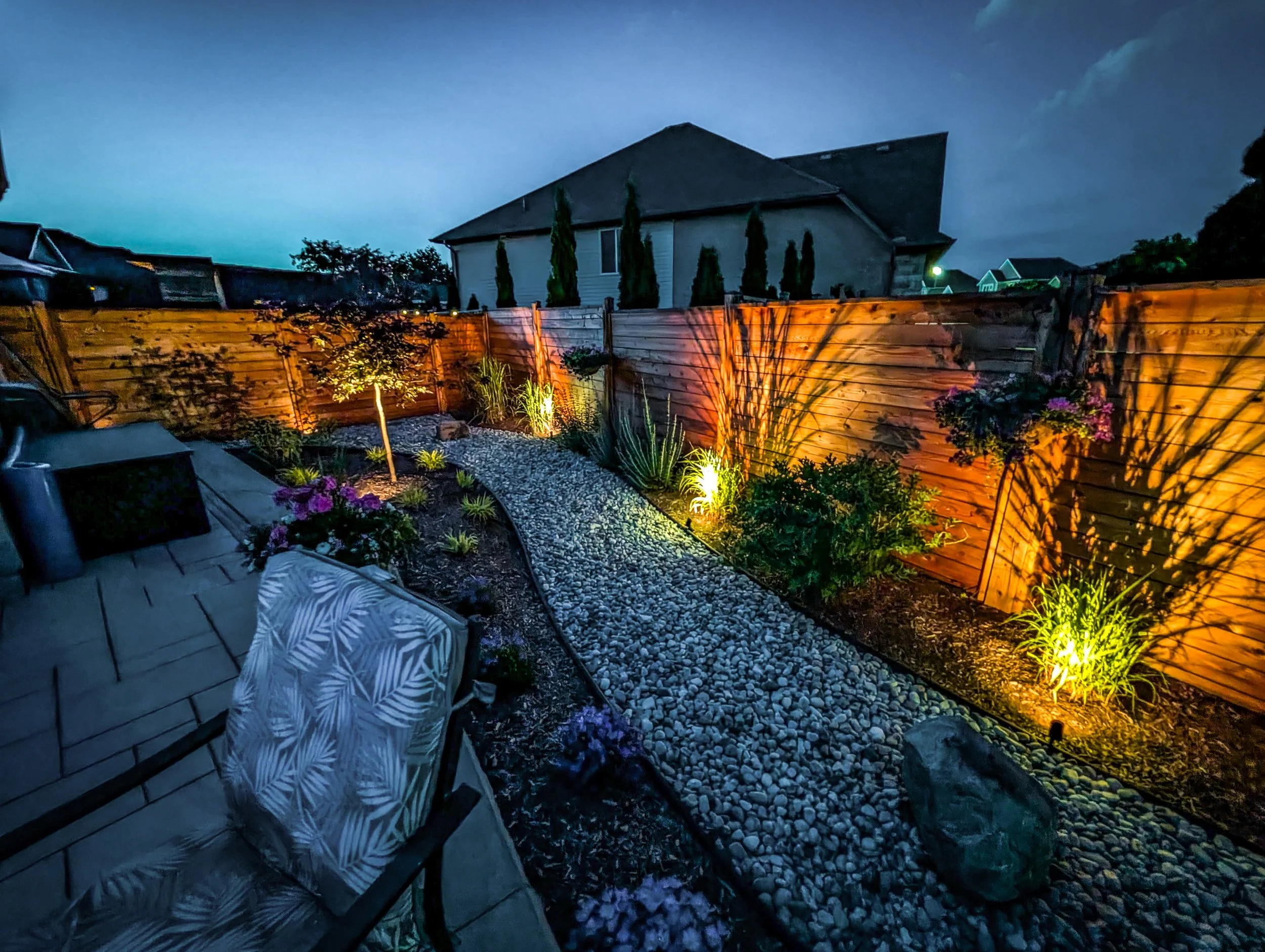 A backyard at night with a wooden privacy fence illuminated by outdoor lighting, a pathway of small rocks, various plants, and a house with a pitched roof in the background under a dark sky.