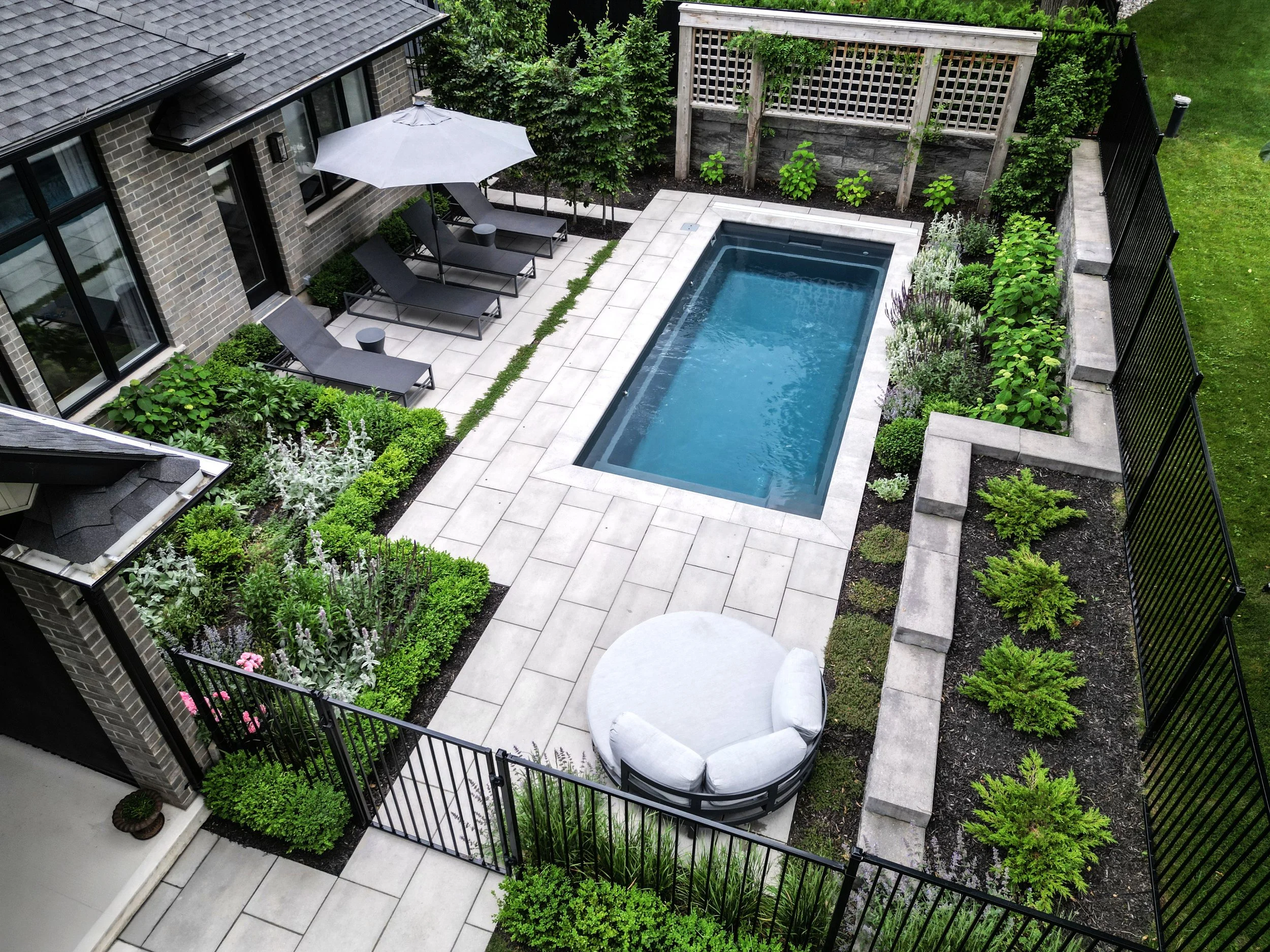 A backyard with a rectangular pool, surrounded by a paved area with lounge chairs and an umbrella, fenced with greenery and shrubs.