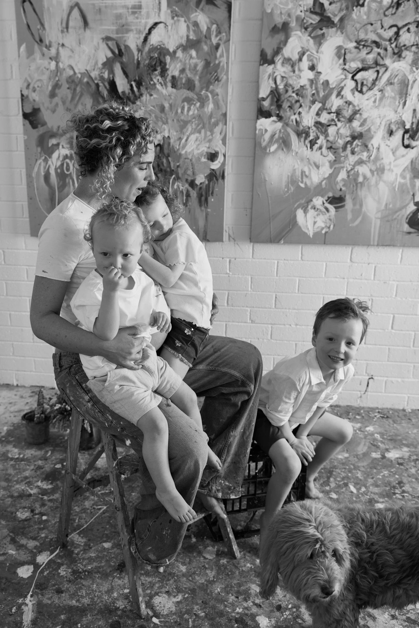 A woman with curly hair holding two children, one on her lap and the other on her shoulder, sitting on a stool in front of an abstract floral painting. A young boy is sitting on a crate nearby, smiling, and a dog is lying on the floor in front of him. The setting appears to be an art studio with a textured floor and a brick wall.