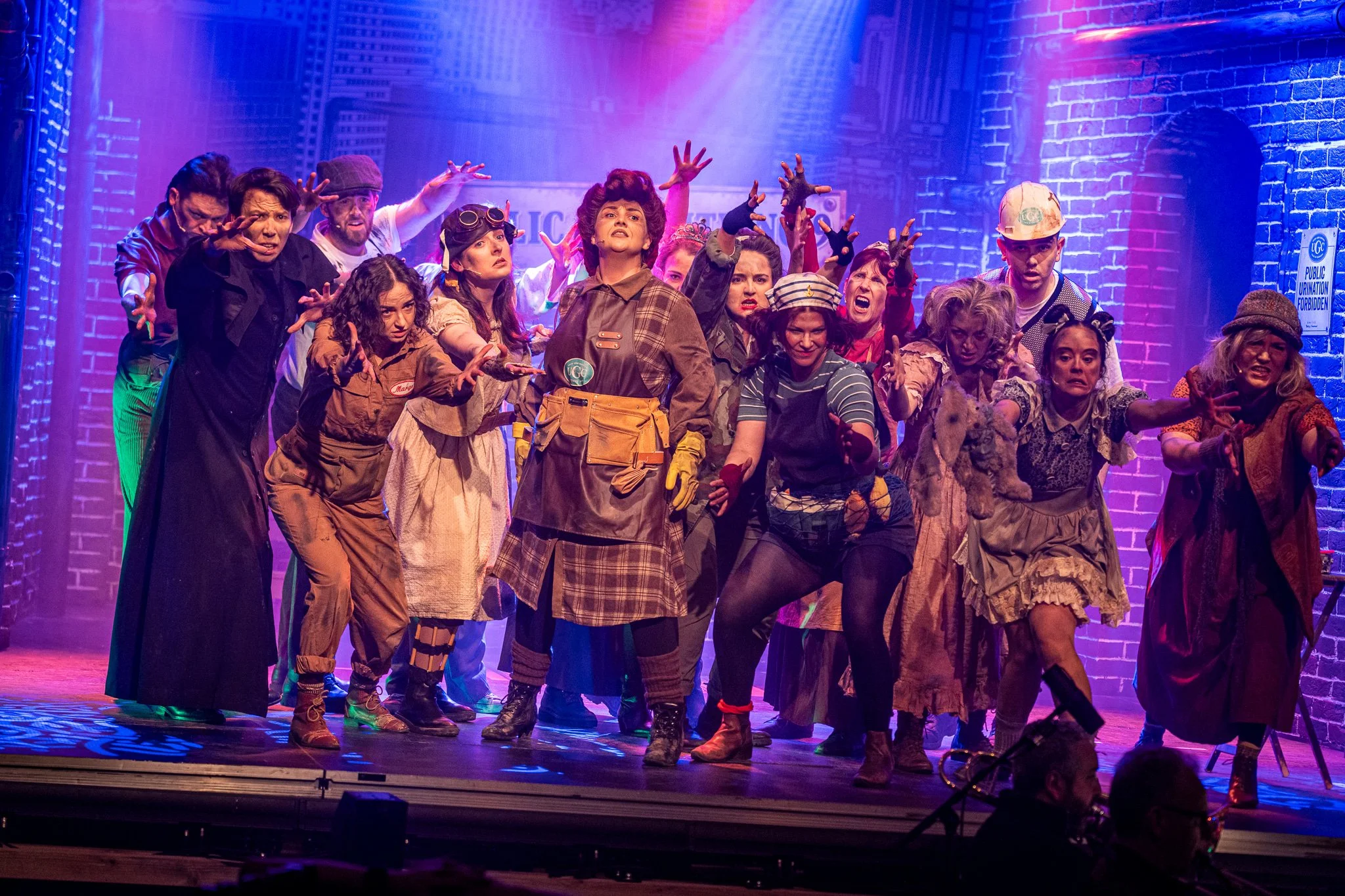 A group of actors on stage in colorful costumes and makeup, performing in a theatrical production with purple and pink lighting and a cityscape backdrop.