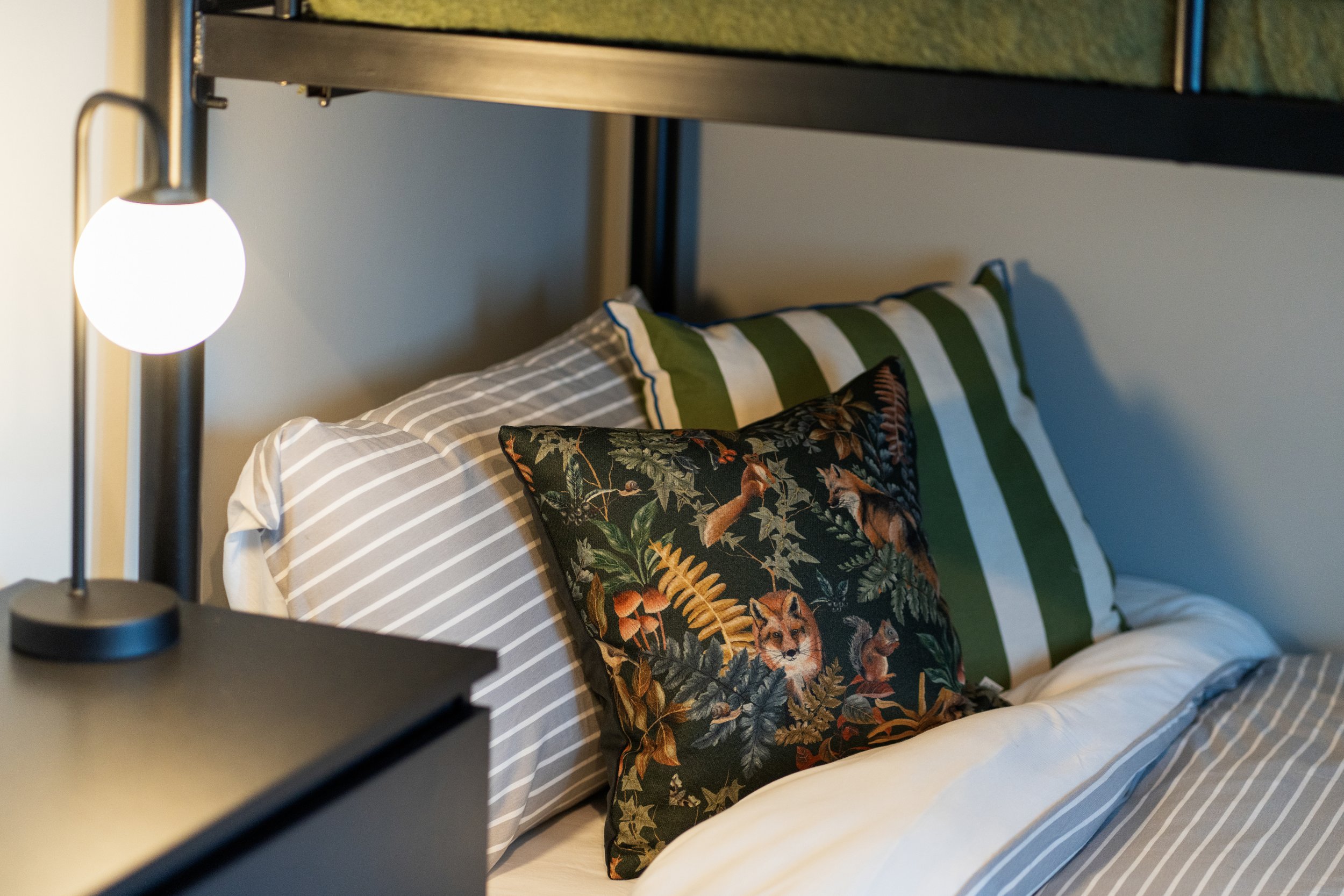Cozy bed with striped bedding and decorative pillow featuring an animal and nature pattern, next to a nightstand with a glowing lamp.