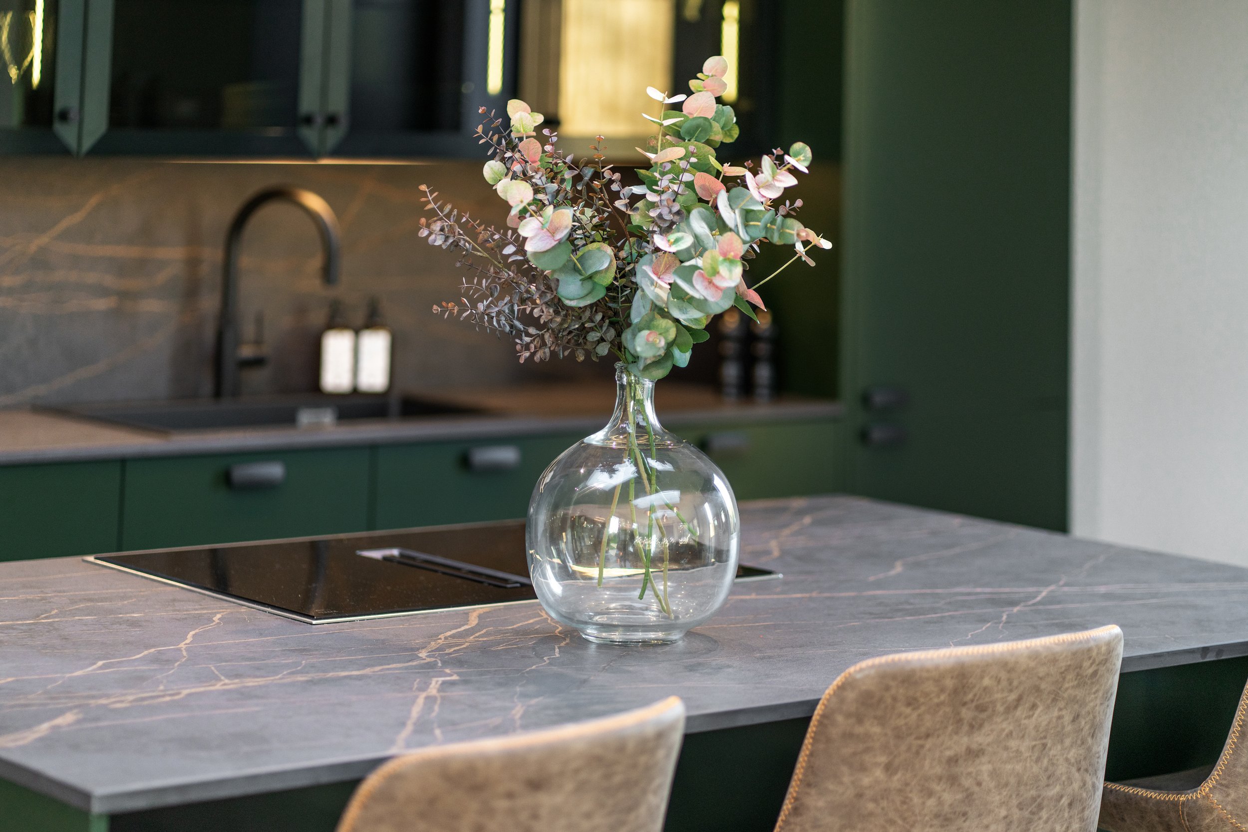 Modern kitchen with marble countertop, glass vase with green and pink eucalyptus branches, dark cabinetry, and sink.