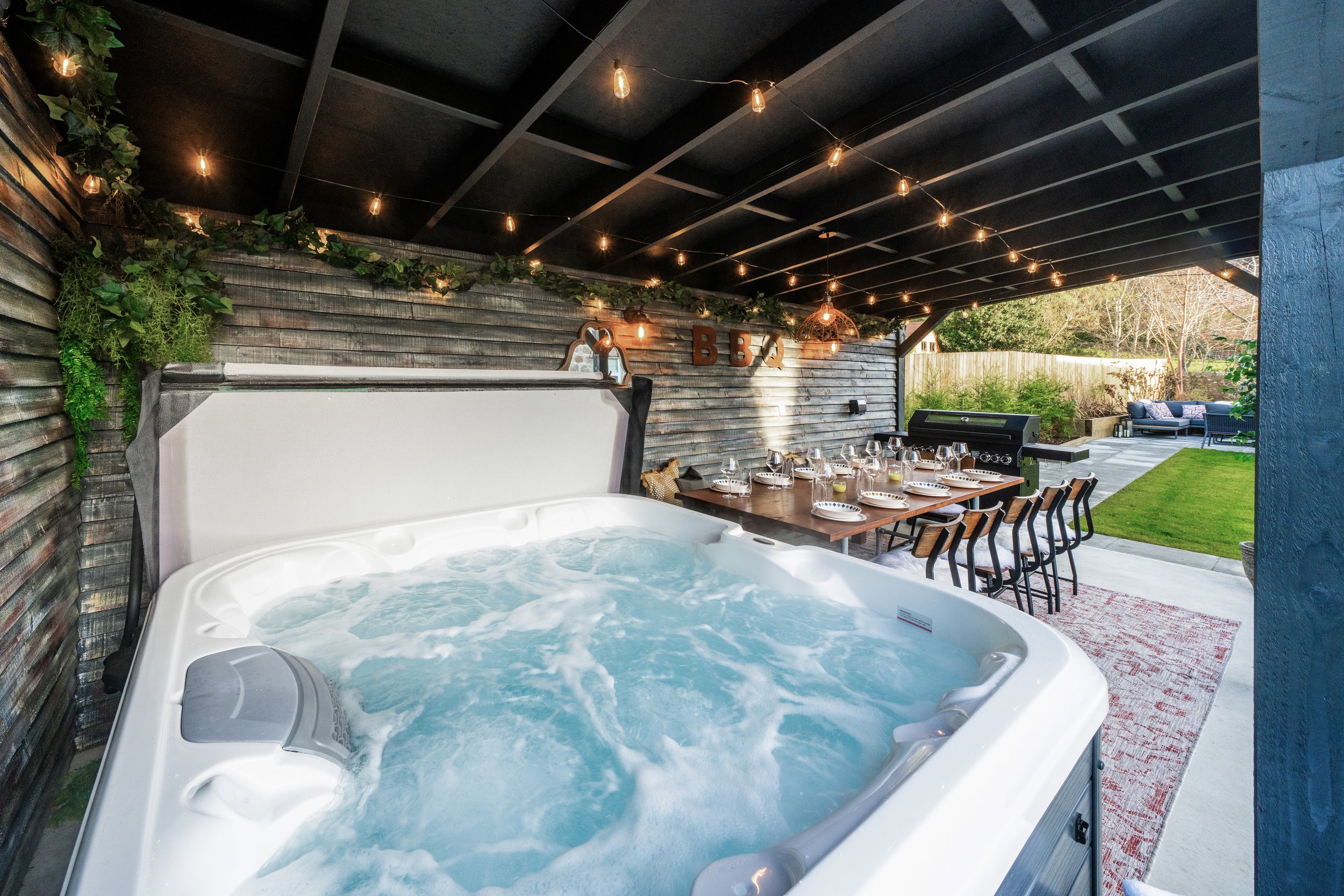 Covered outdoor area with hot tub, dining table set for a meal, barbecue grill, and string lights, surrounded by greenery.