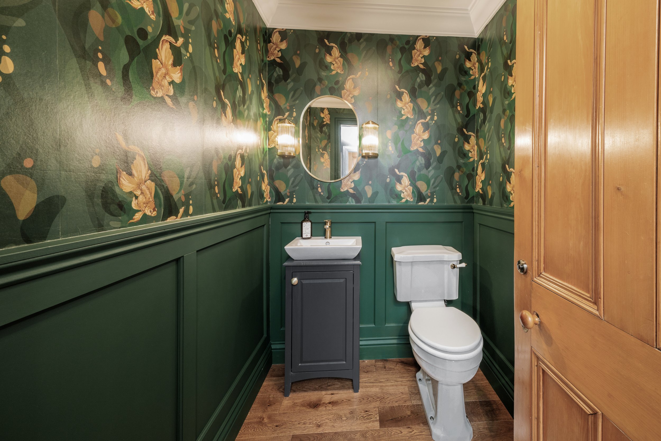 Bathroom with floral wallpaper, a round mirror, a small sink with a cabinet, a toilet, wooden flooring, and wood paneling.
