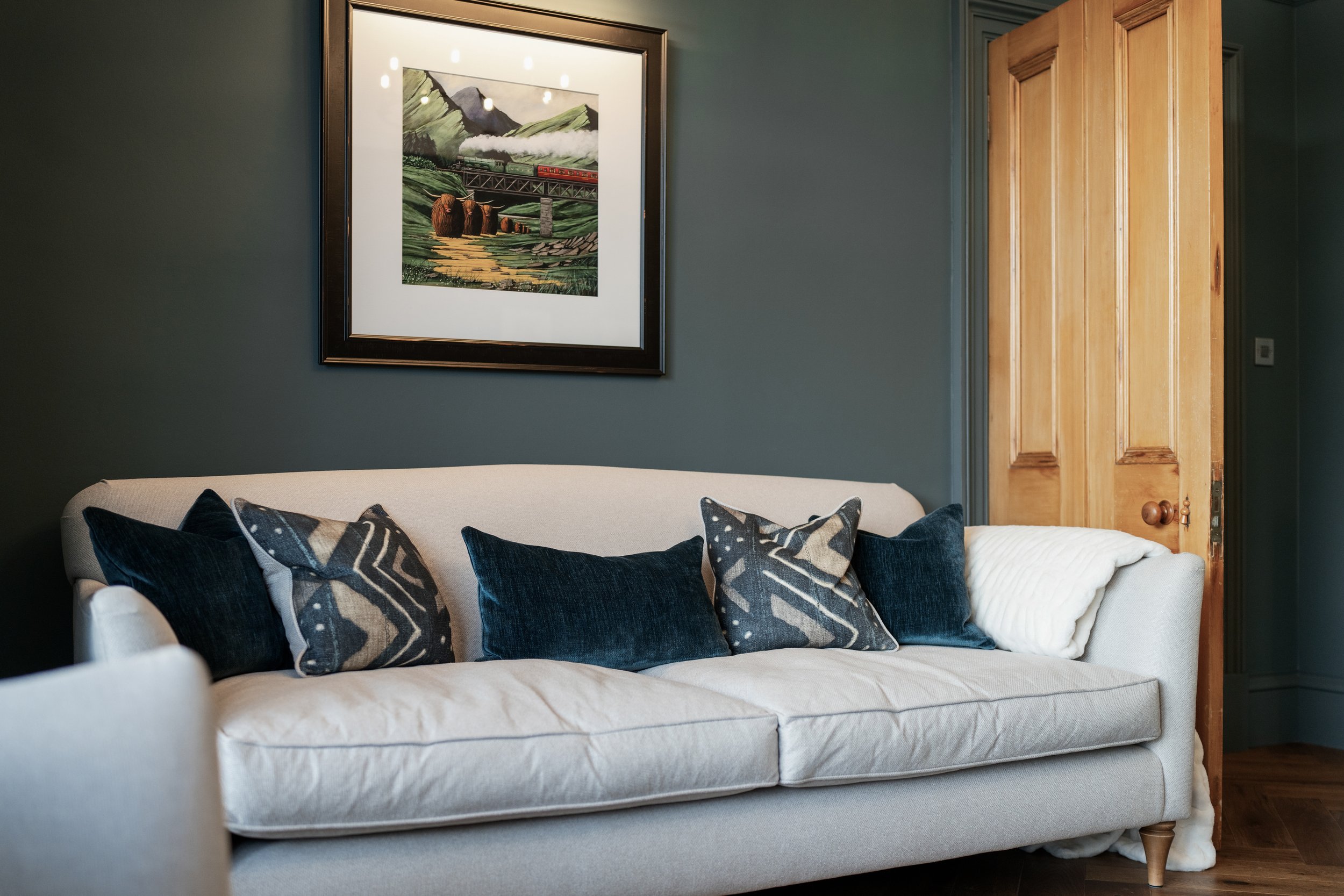 Living room with beige sofa, blue and patterned pillows, framed artwork, and an open wooden door.