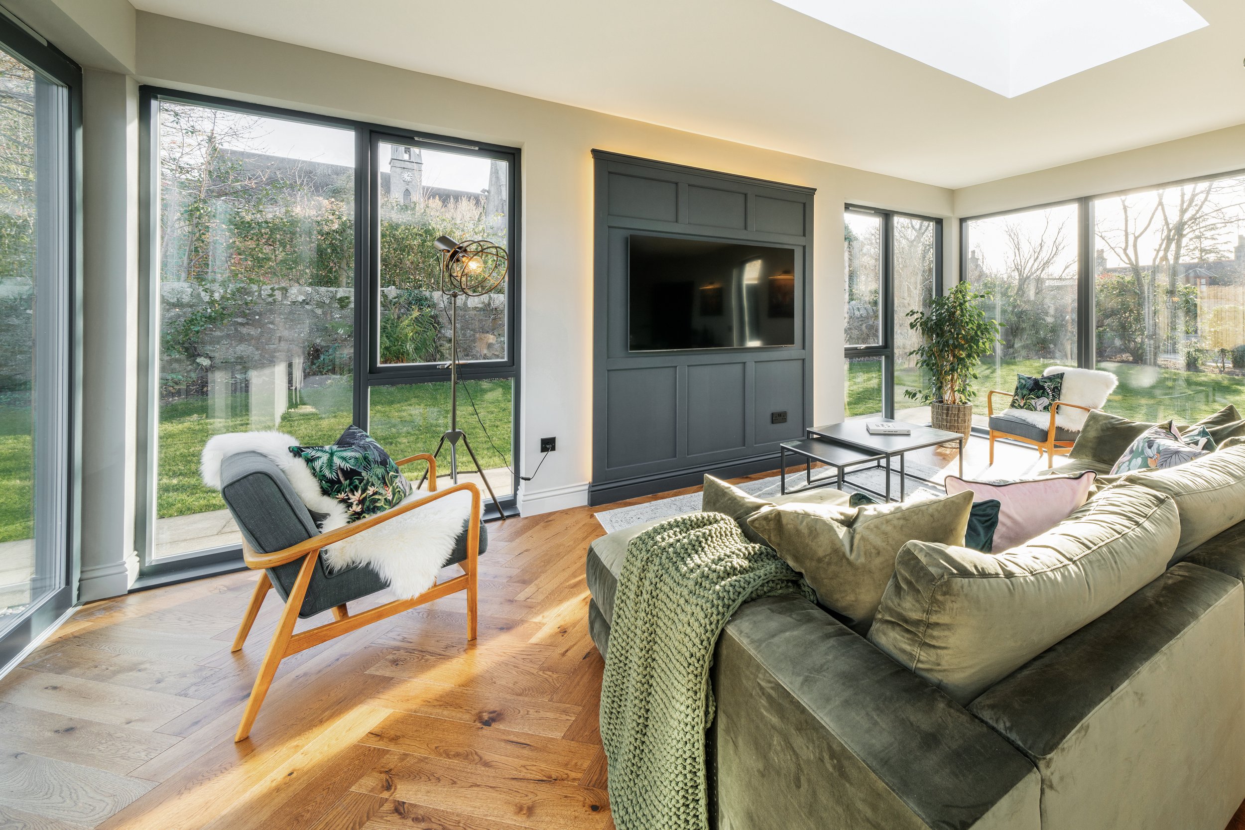 Modern living room with large windows, featuring a green sofa, wooden chairs, a wall-mounted TV, and hardwood floors. The room is well-lit with natural light, offering a view of the garden outside.