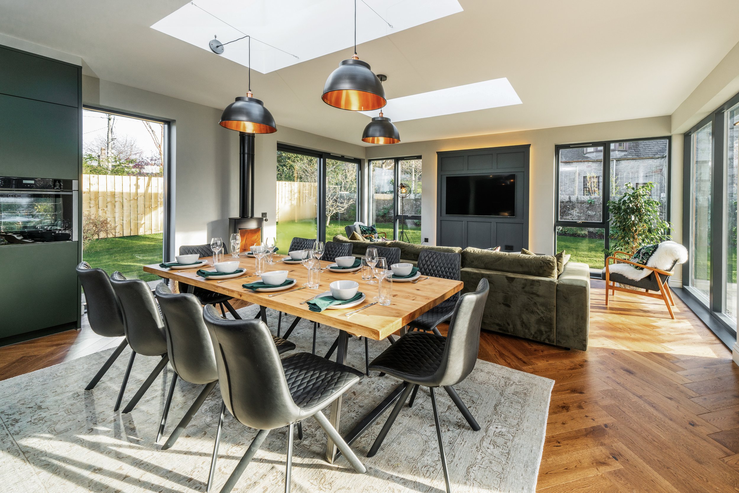 Modern dining and living area with large windows, dining table set with plates and glasses, black chairs, green sofa, indoor plant, and a wood stove.
