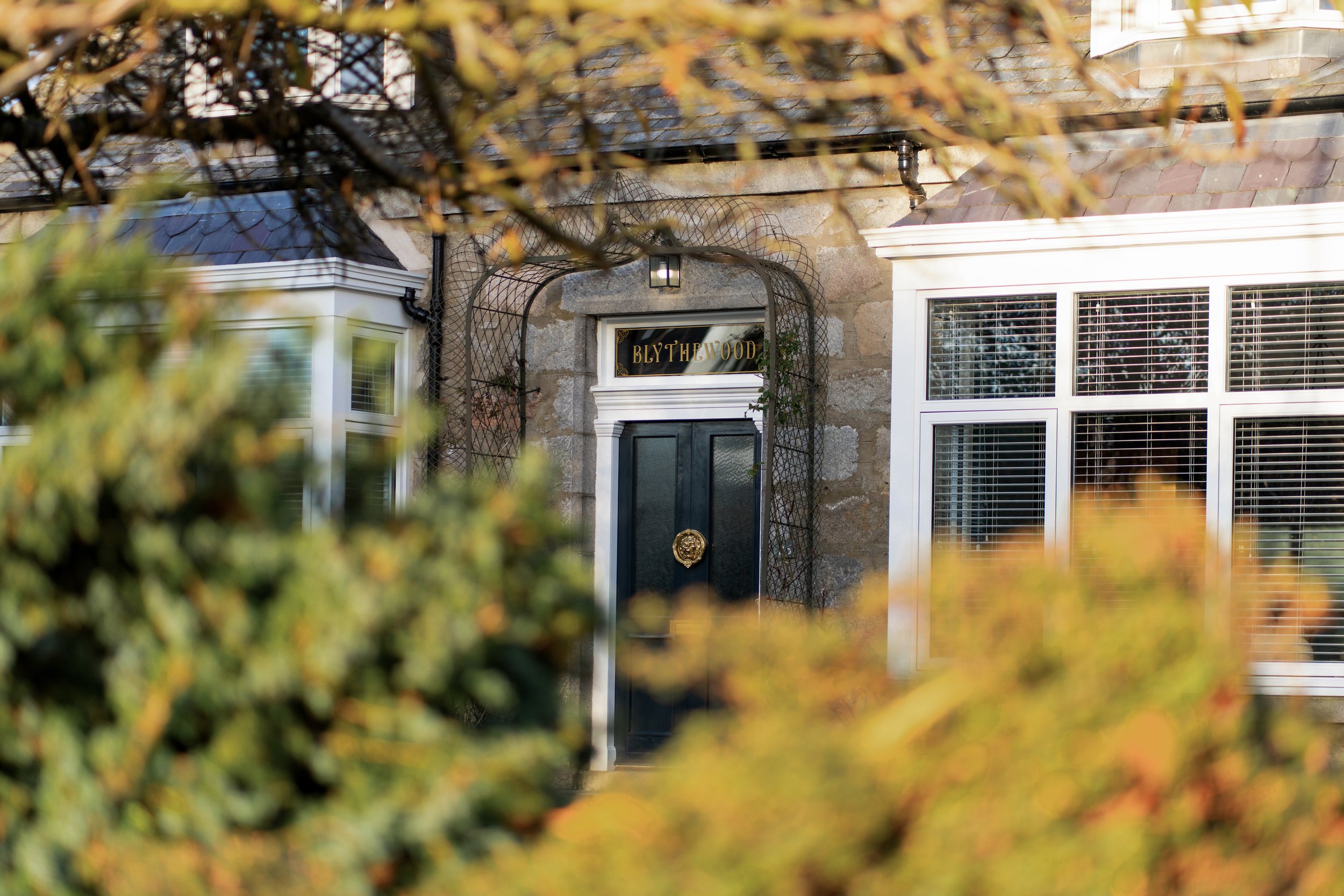 Front entrance of house with dark door, stone walls, large windows, and greenery in foreground.