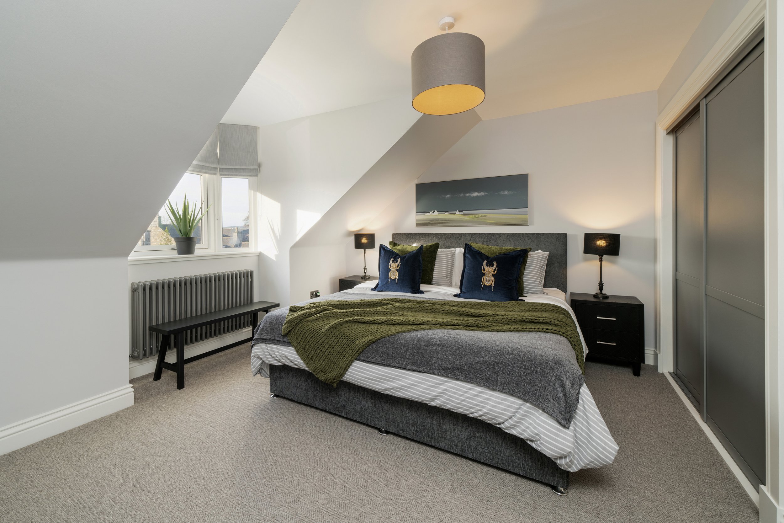 Modern bedroom with gray and green decor, double bed with pillows and throws, two bedside tables with lamps, a wall-mounted painting, a small potted plant on the window sill, and a bench under the window.
