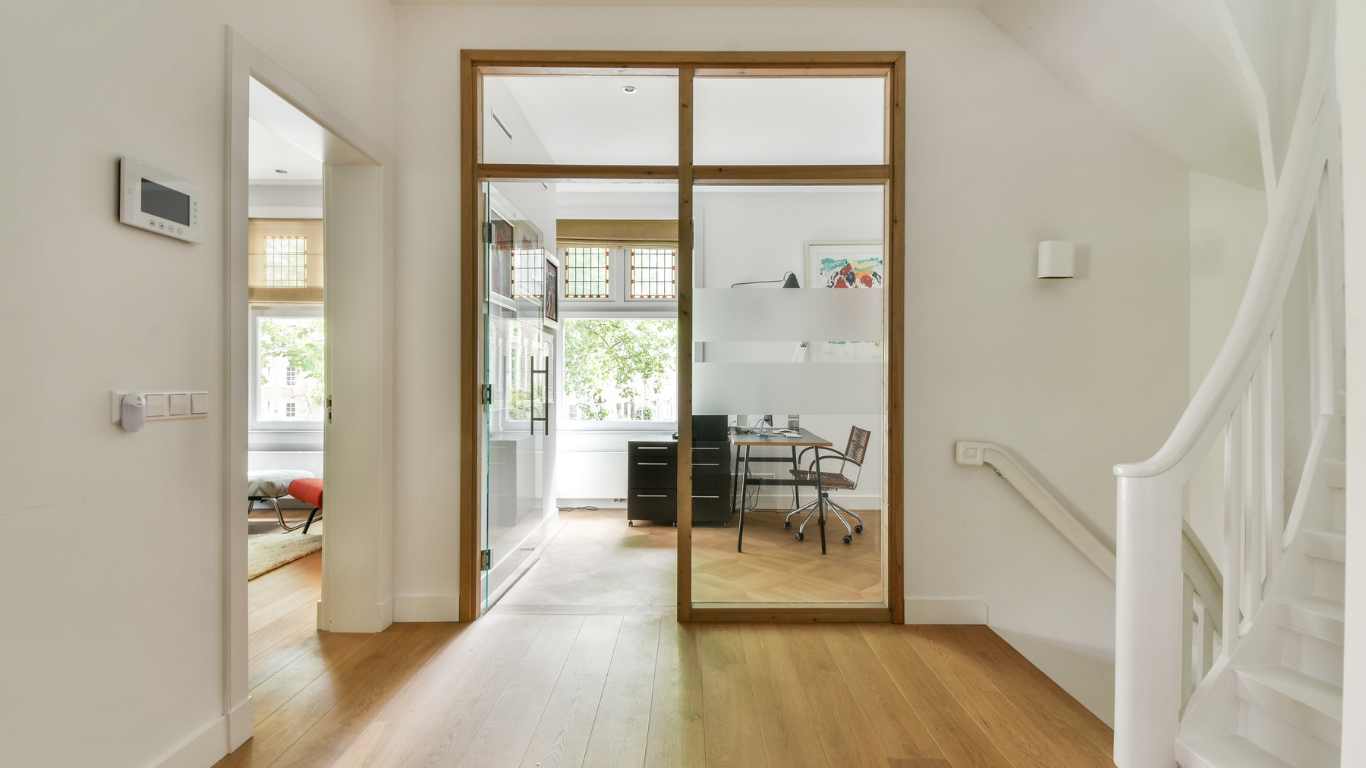 Interieur van een woning met een lichte hal, een aparte werkruimte gescheiden door een glazen deur met houten frame, een trap rechts, en een kamer links met groot raam en rood-bank.