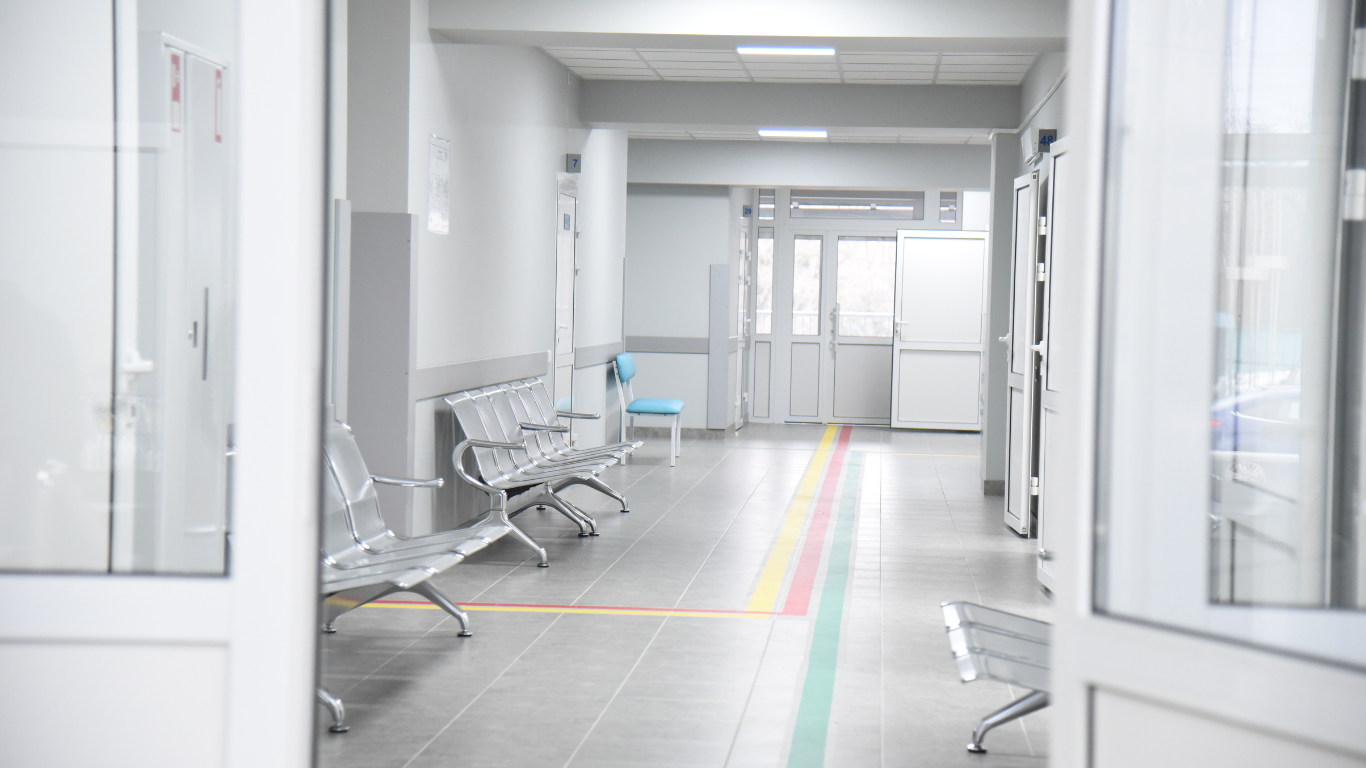 Lege wachtkamer in een koelkamer of kliniek met rijen lege metalen stoelen, witte muren, een deur en ramen in de achtergrond.