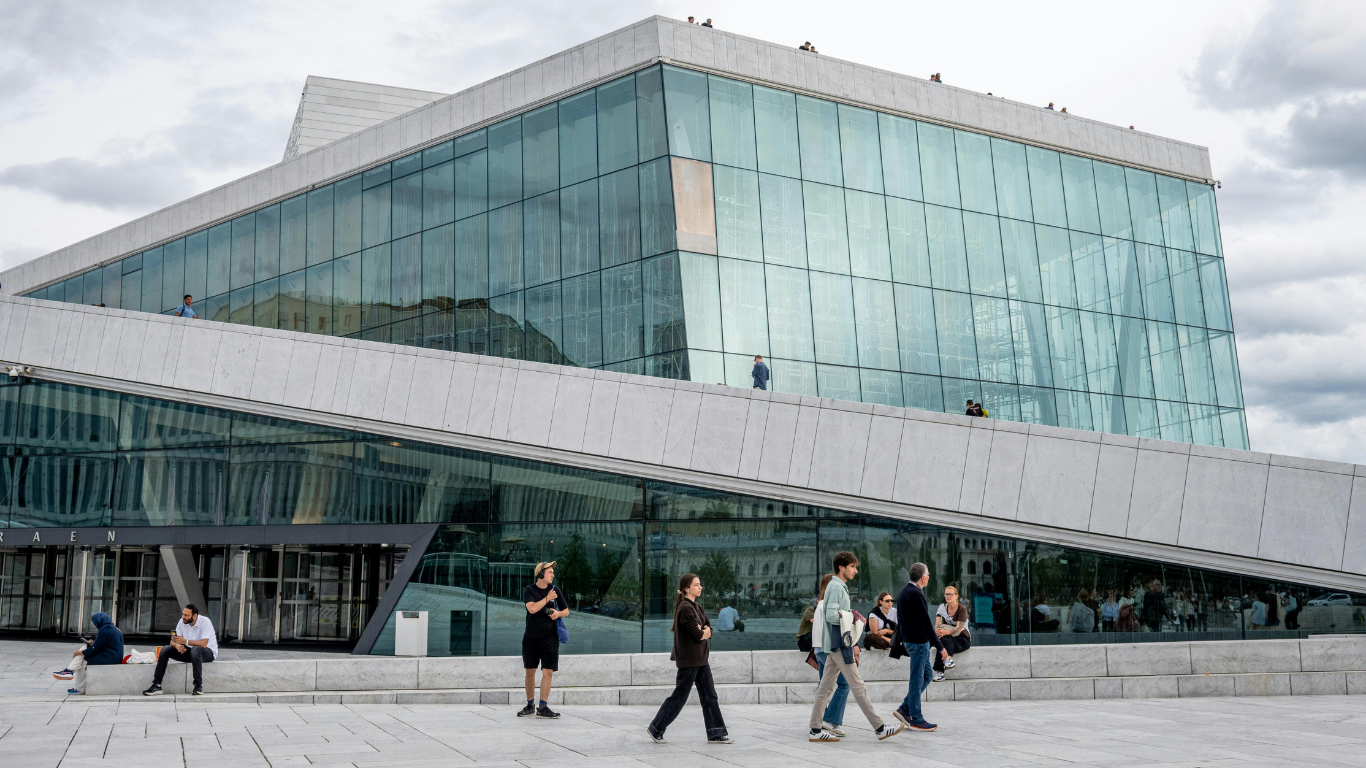 Modern gebouw met grote glazen ramen en een hellend terras, met mensen die eroverheen lopen en rusten, onder een bewolkte lucht.