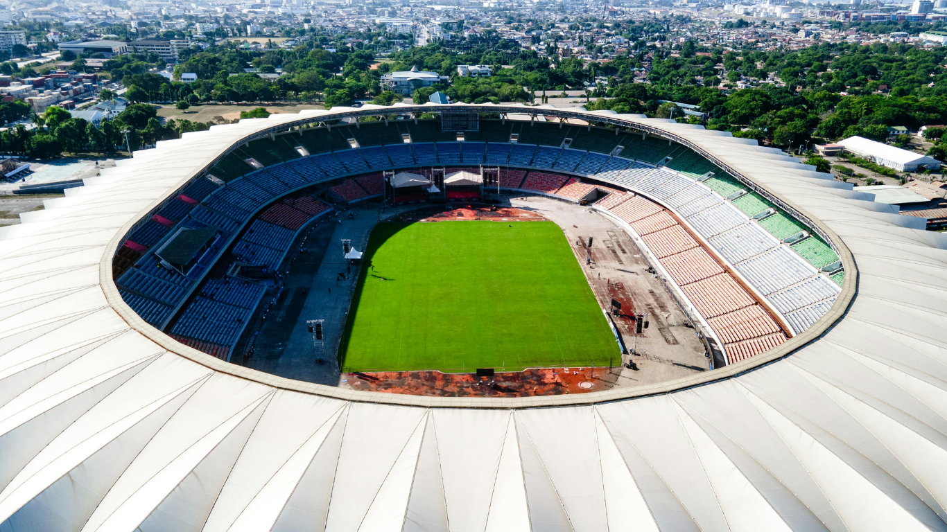 Luchtfoto van een voetbalstadion met groen veld, omringd door tribunes en een moderne architectuur, in een stedelijke omgeving.
