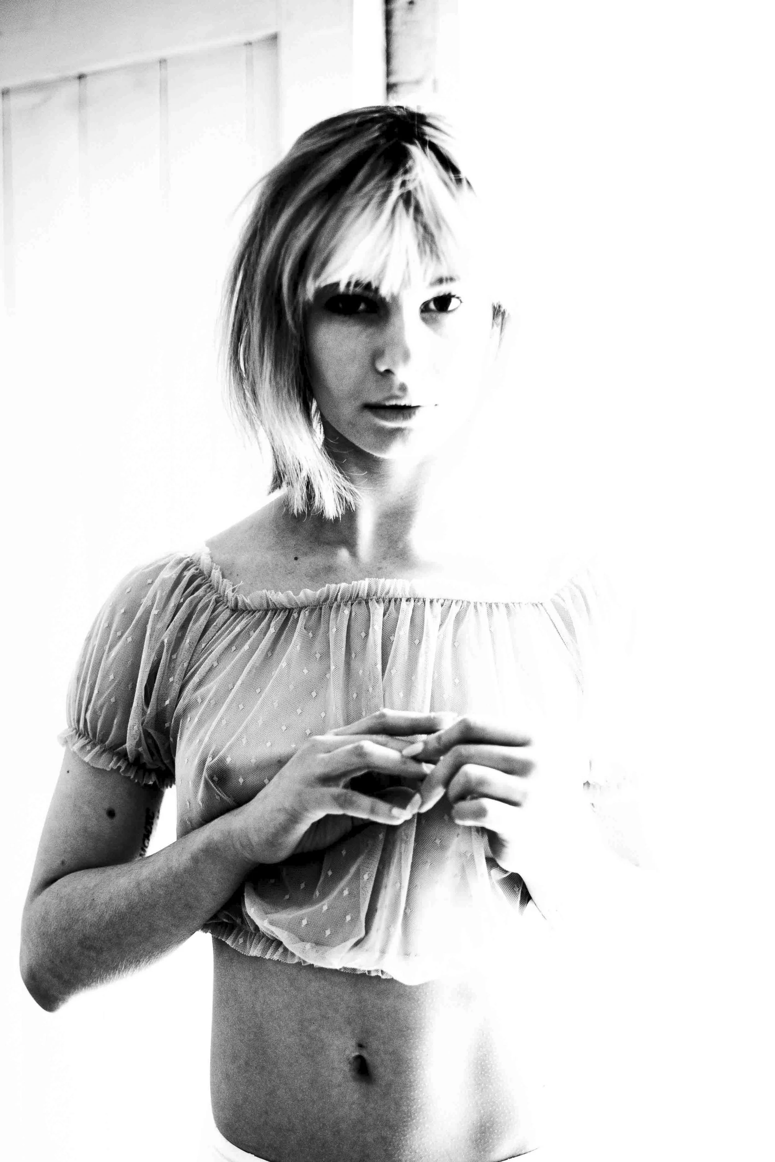 Black and white photo of a young woman with short hair holding a glass, wearing a light-colored ruffled top, standing against a plain background.