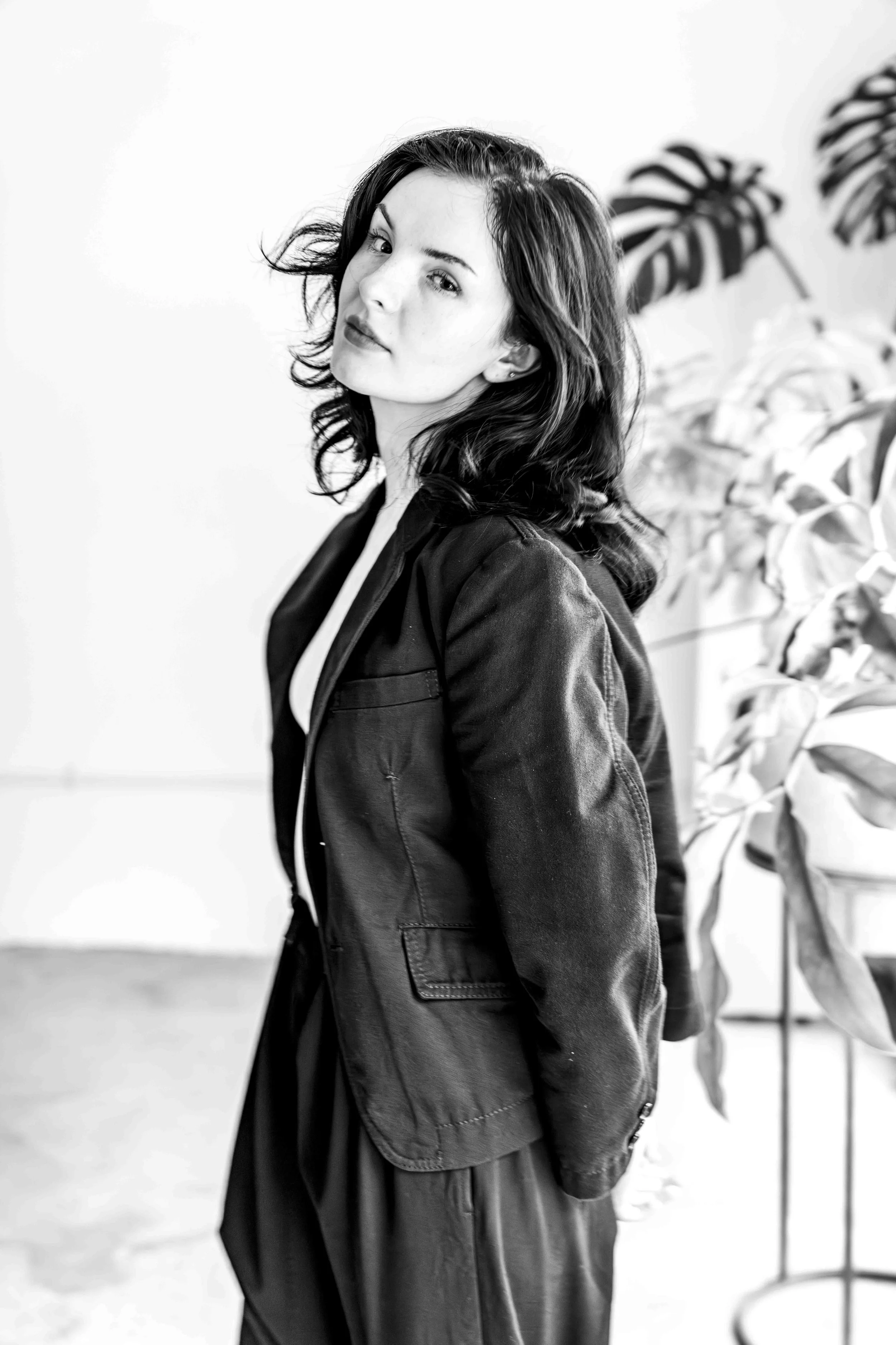 Black and white portrait of a woman with dark wavy hair, wearing a black leather jacket, standing near plants on a white background.