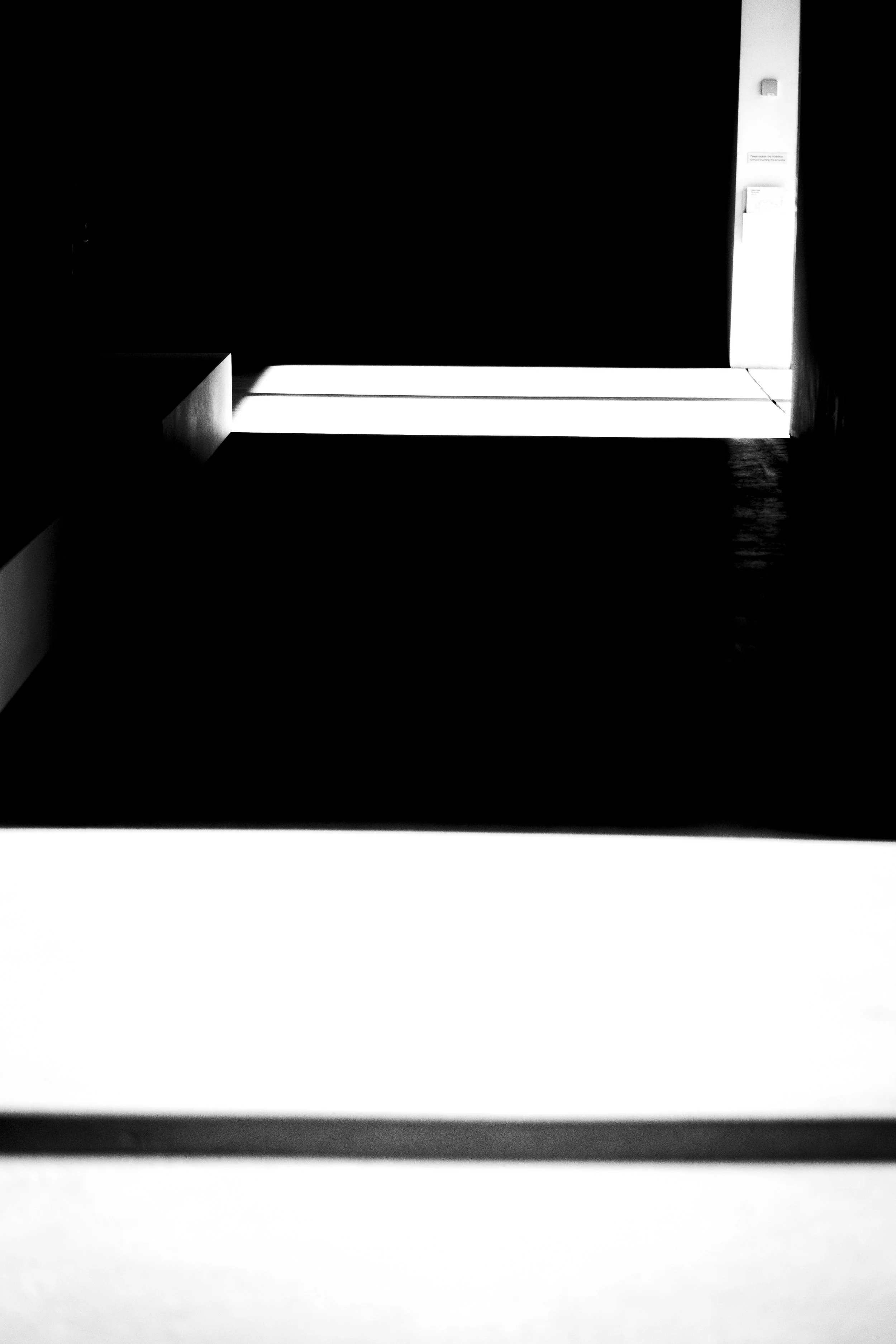 Black and white photograph of a minimalistic, modern interior space with a large rectangular window or opening, allowing natural light to cast sharp shadows on the floor and walls.