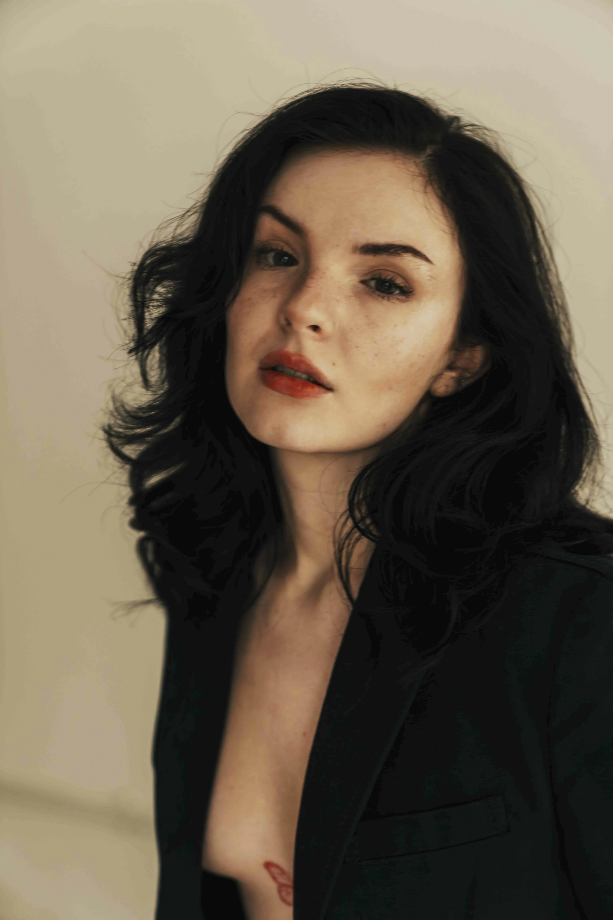 Close-up of a woman with black, wavy hair, light skin, and blue eyes, wearing a black top and displaying a small red tattoo butterfly on her chest, against a plain beige background.