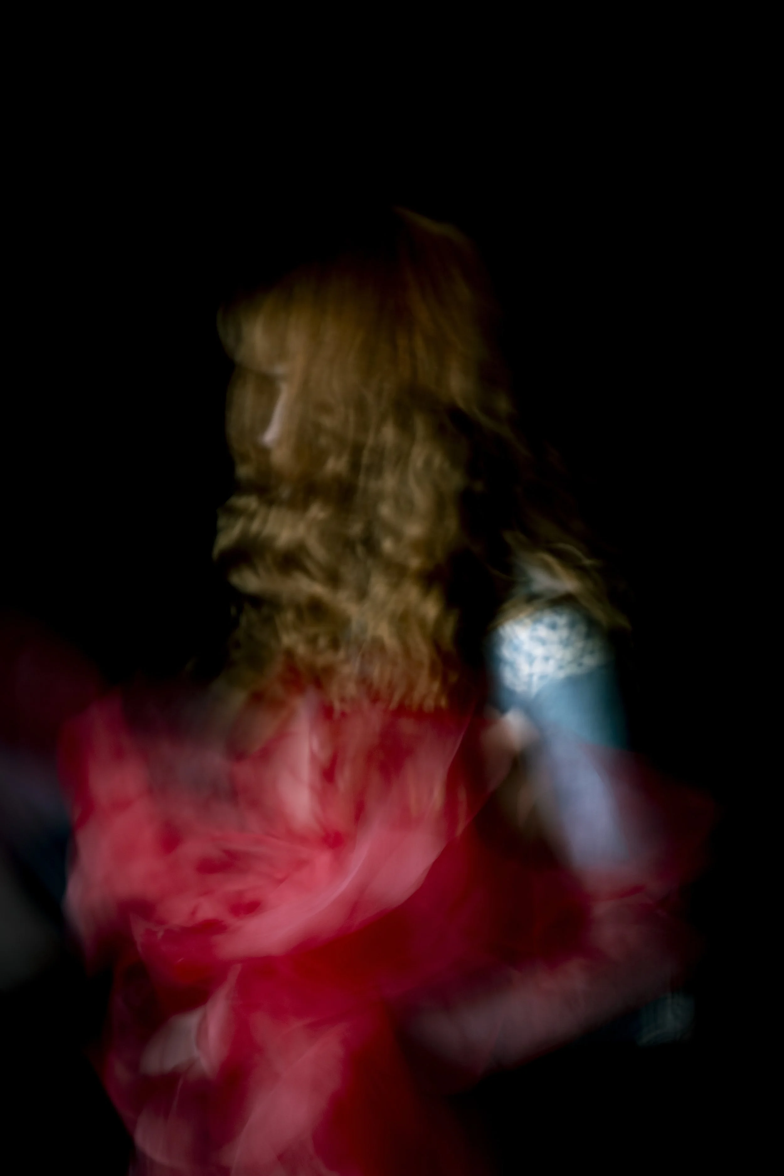 A woman with long curly brown hair looking to the side, wearing colorful clothing with a dark background.